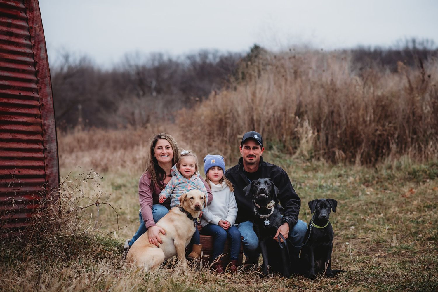 Family photo session outdoors with three dogs against rustic red barn and tall grass backdrop.