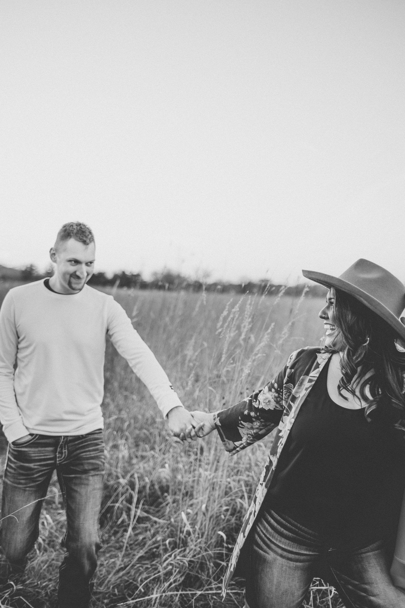 A black and white photo captures a couple holding hands and laughing in a meadow.