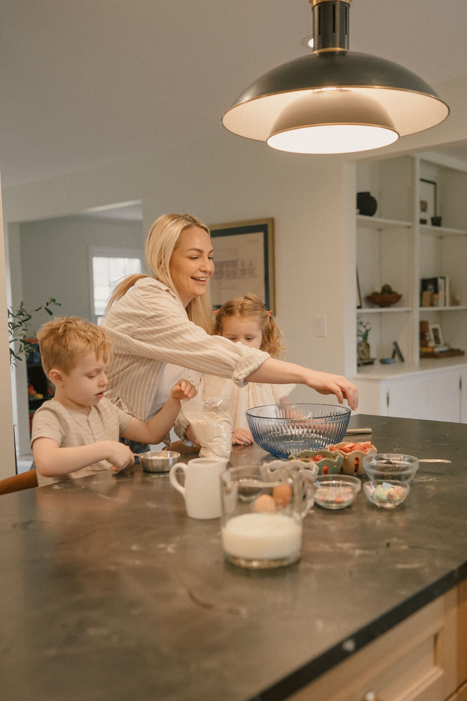 A smiling mother bakes with her two young children at a kitchen island with ingredients and bowls.