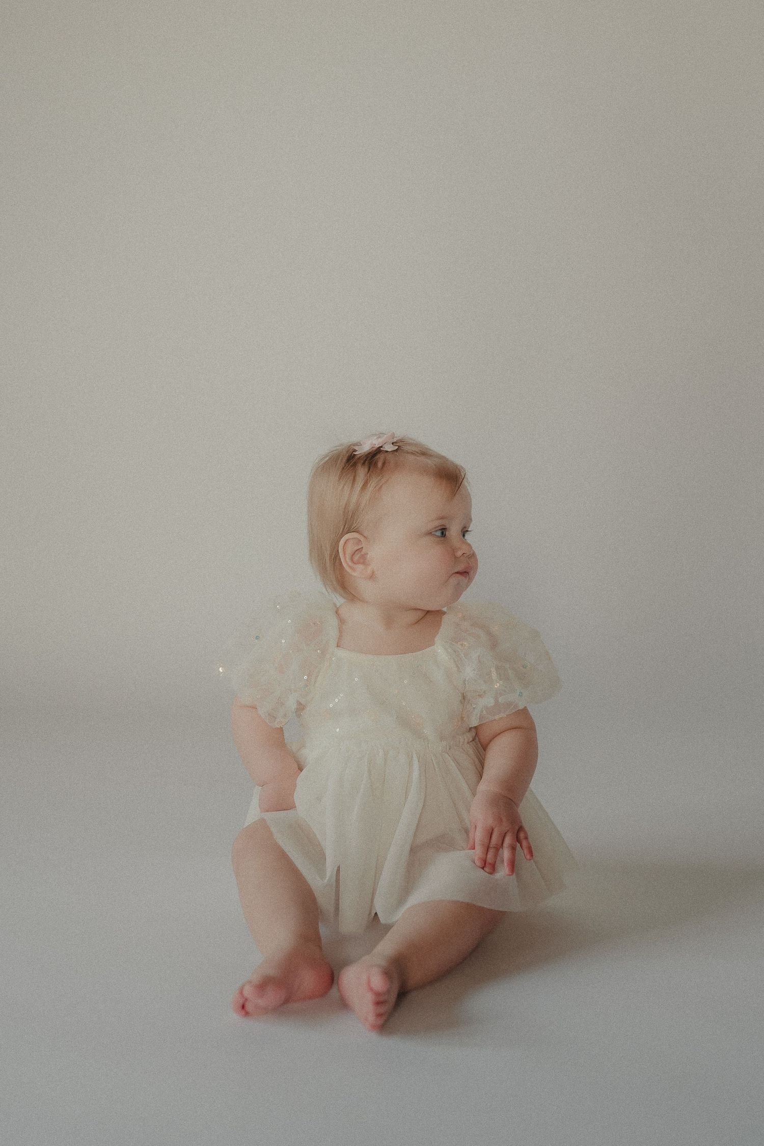 Sweet baby girl in a white tulle dress sits barefoot on a light background during her one year photo session.