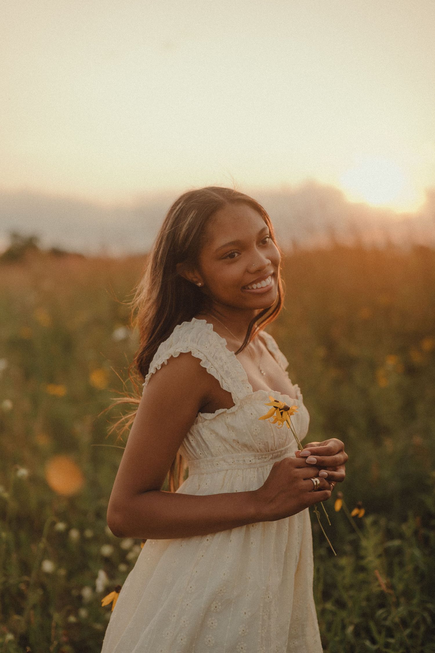 Smiling young woman in white dress holds yellow wildflower in a sunlit field at golden sunset.