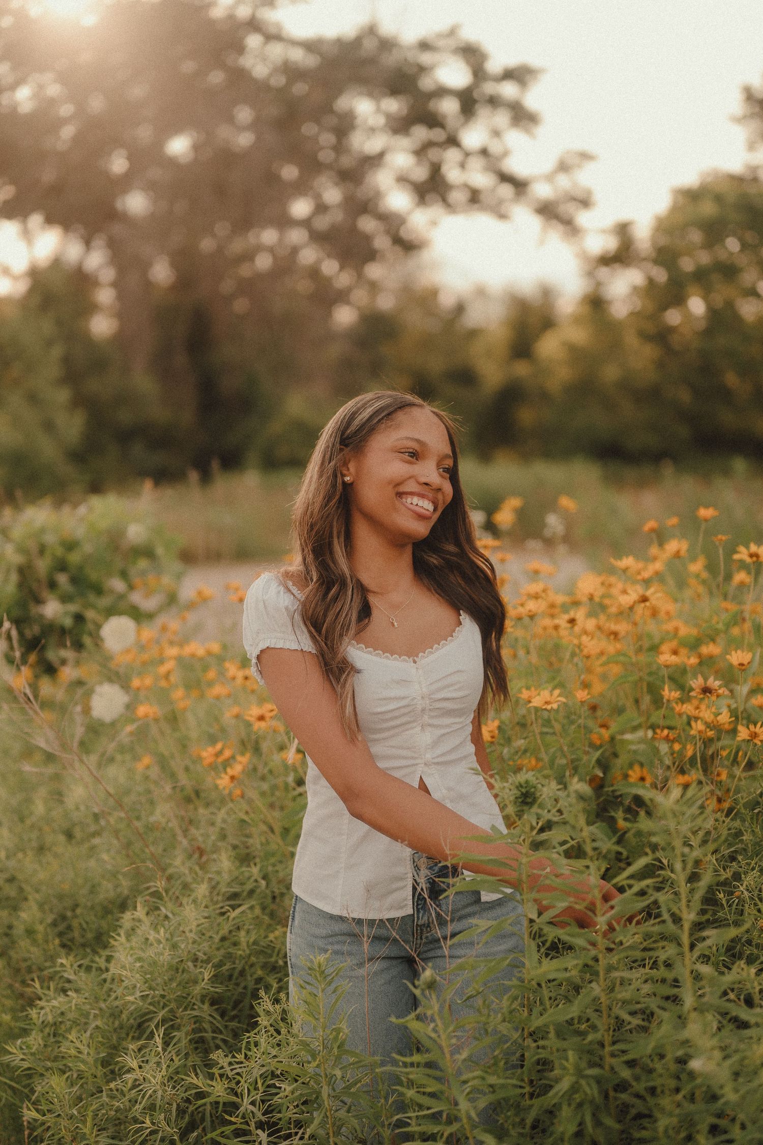 Young woman smiling in a field of yellow wildflowers at golden hour, wearing white top and jeans.