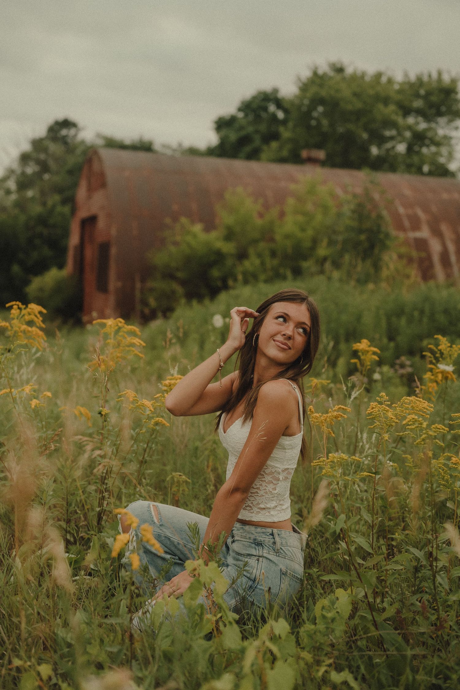 Young woman in white lace top and ripped jeans sitting in a field near a rustic red barn.