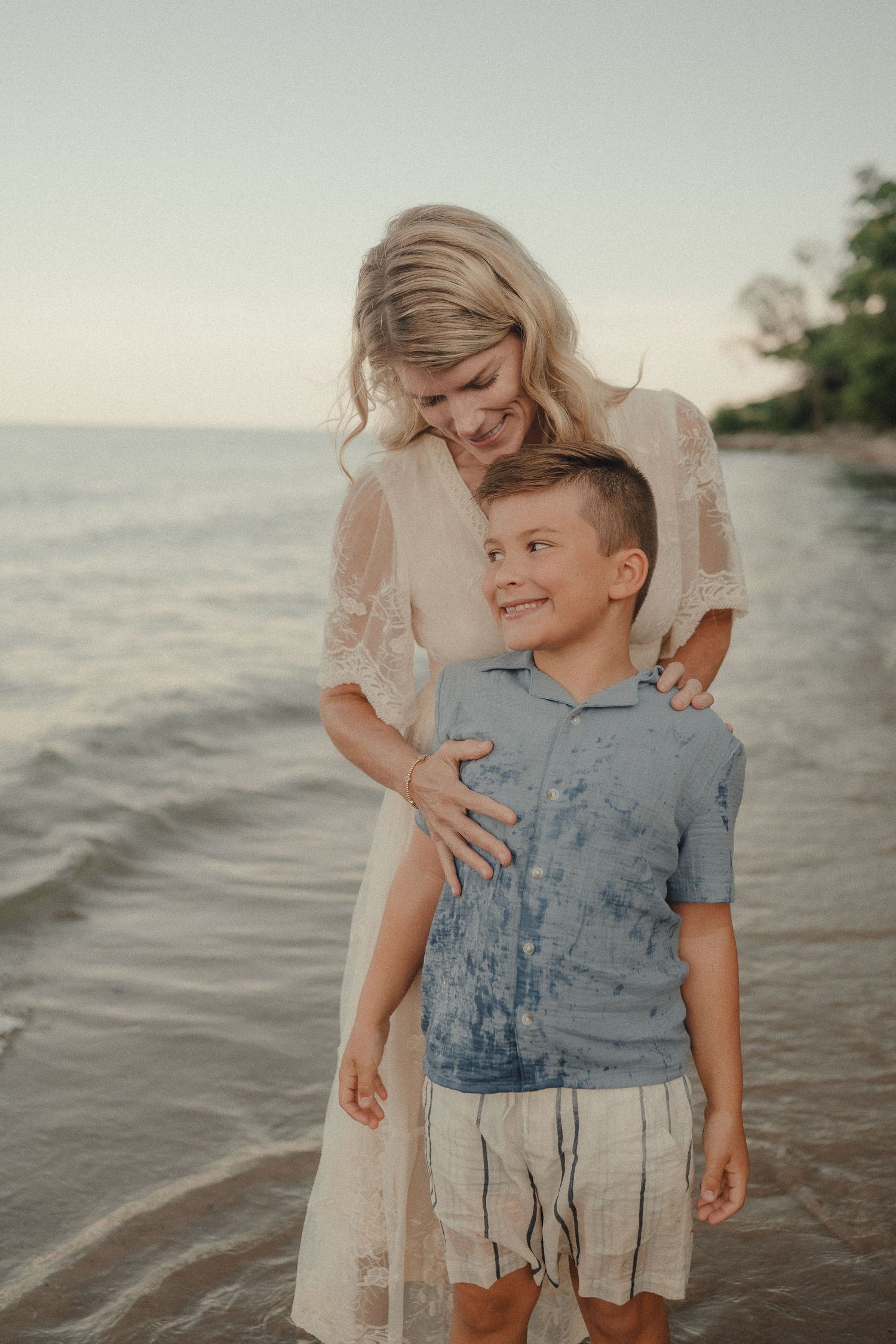 Mother in white lace dress hugging her smiling son while standing in gentle ocean waves.