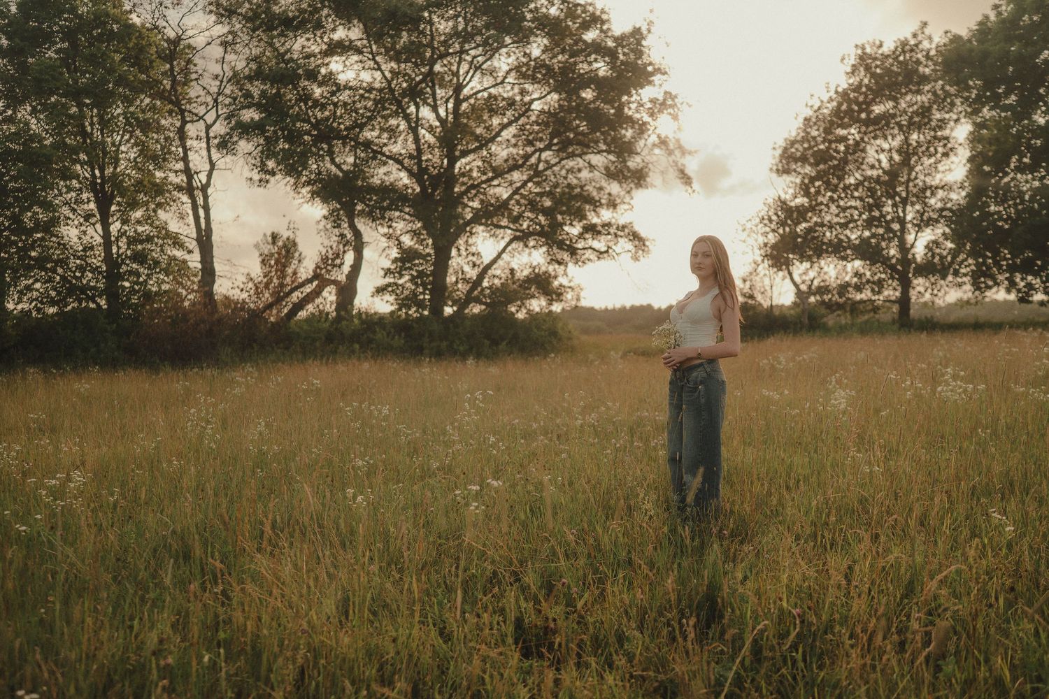 Young woman standing in a sunlit meadow with wildflowers and trees at golden hour.