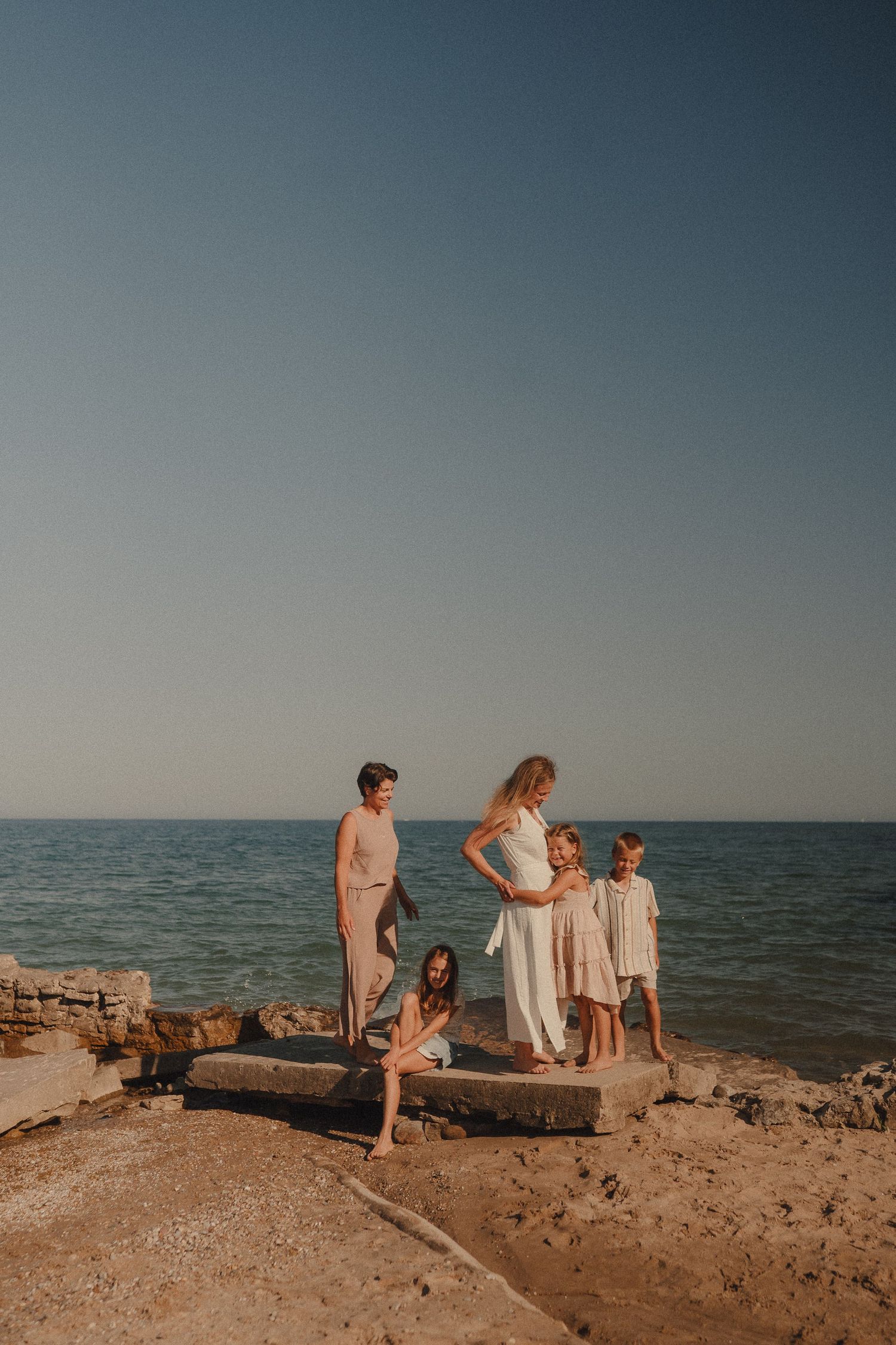 A family of five poses on rocky seaside platform with clear blue sky and ocean in background.