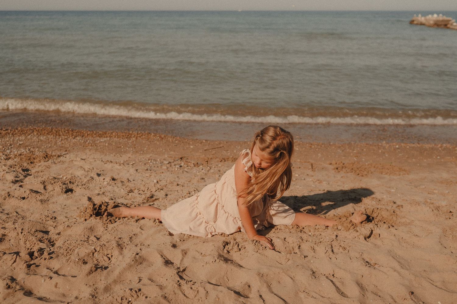 Young girl in white dress playing in sand on a sunny beach near the water.