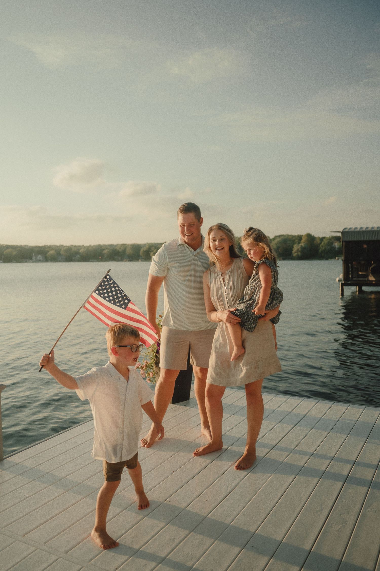 Joyful family with boy waving American flag on white dock at lake sunset.