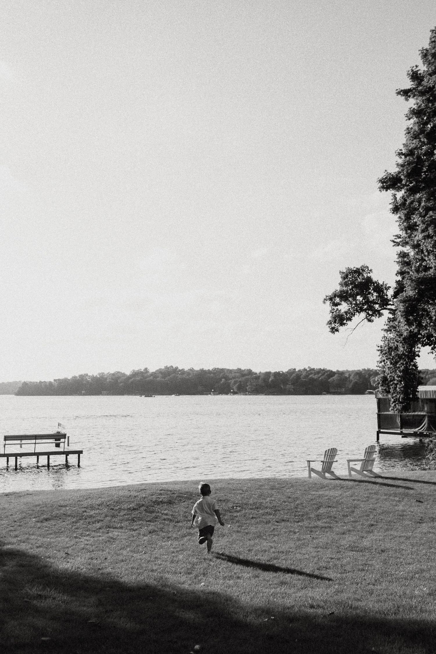 Child running toward a lake with dock and Adirondack chairs in black and white photo.