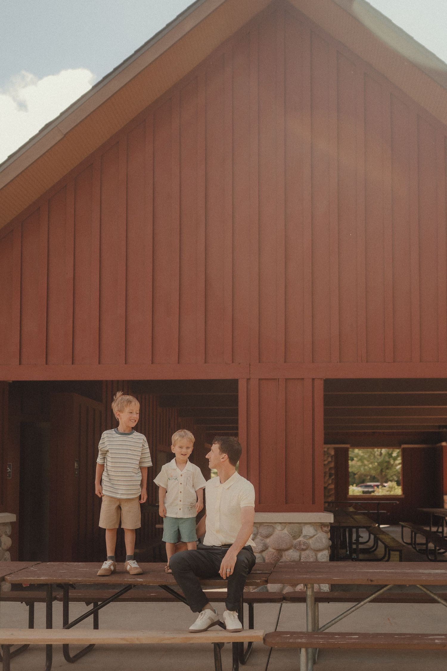 A father sits on a picnic table chatting with his two young sons in front of a red barn.