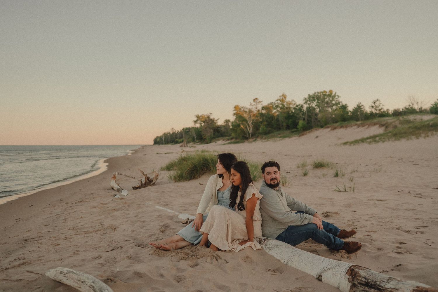 Couple sitting together on sandy beach at sunset with dunes and trees in background.