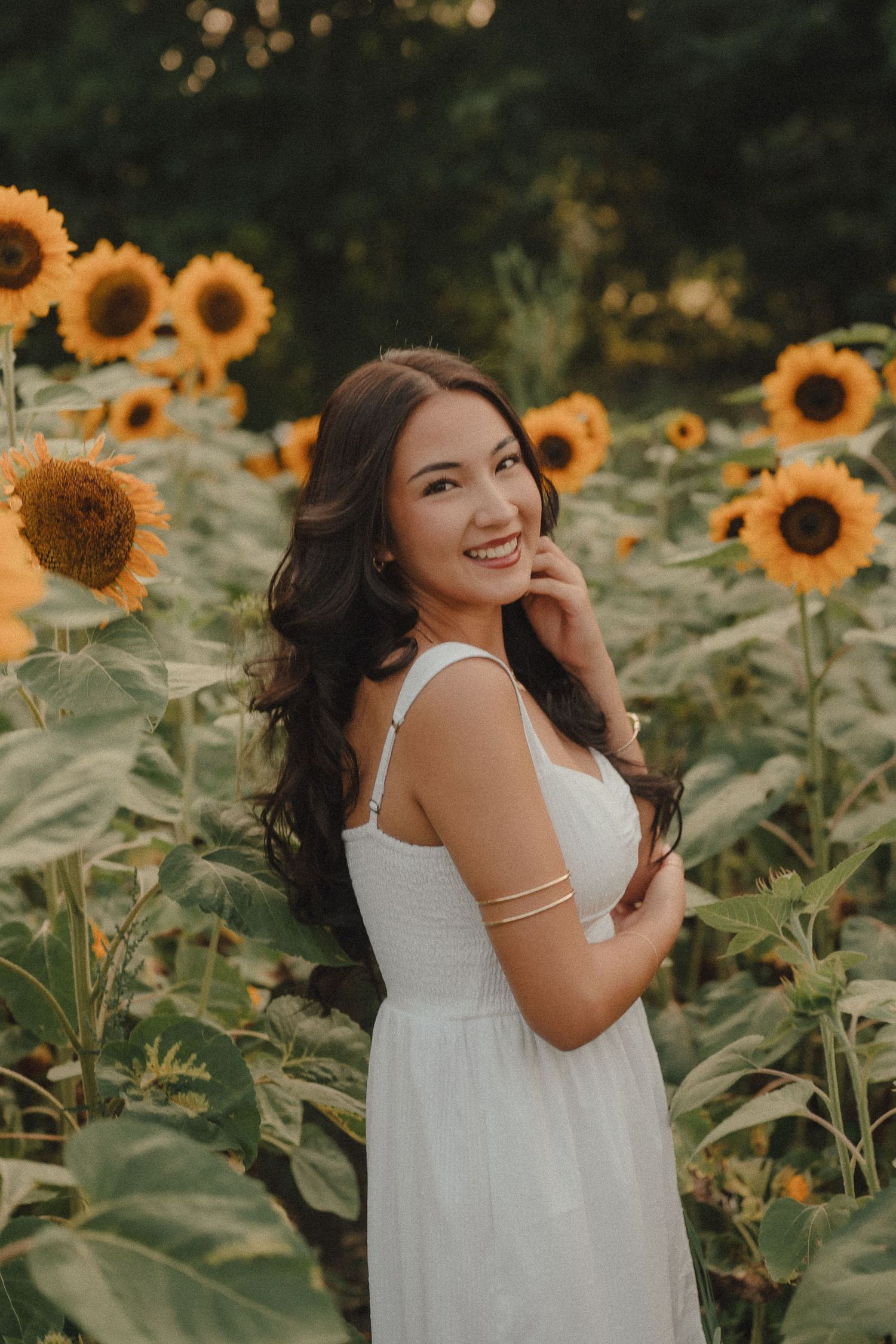 Young woman in white dress smiling among sunflowers at golden hour.