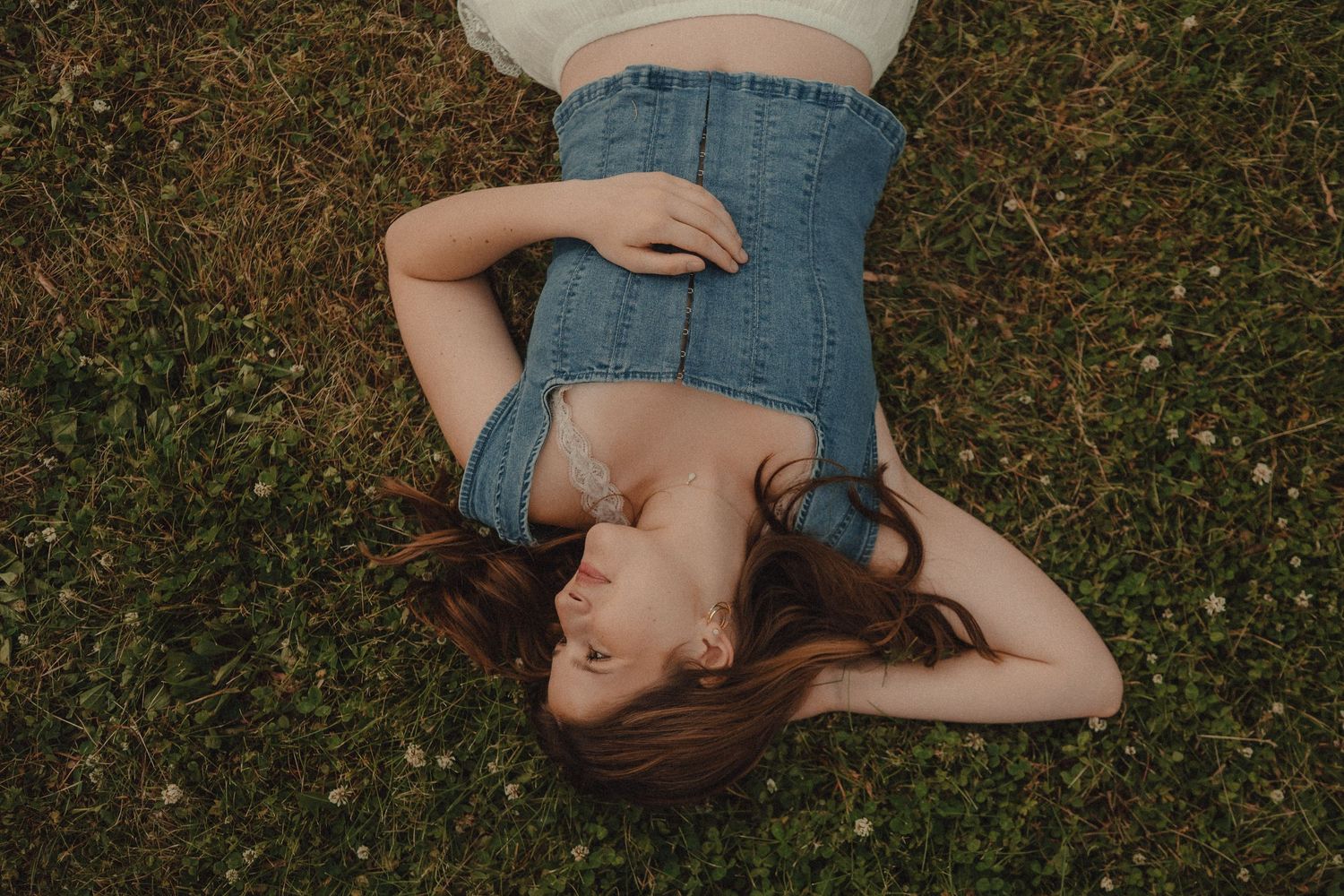 Young woman with long hair lying on green grass, wearing a white crop top and denim skirt, relaxing outdoors.