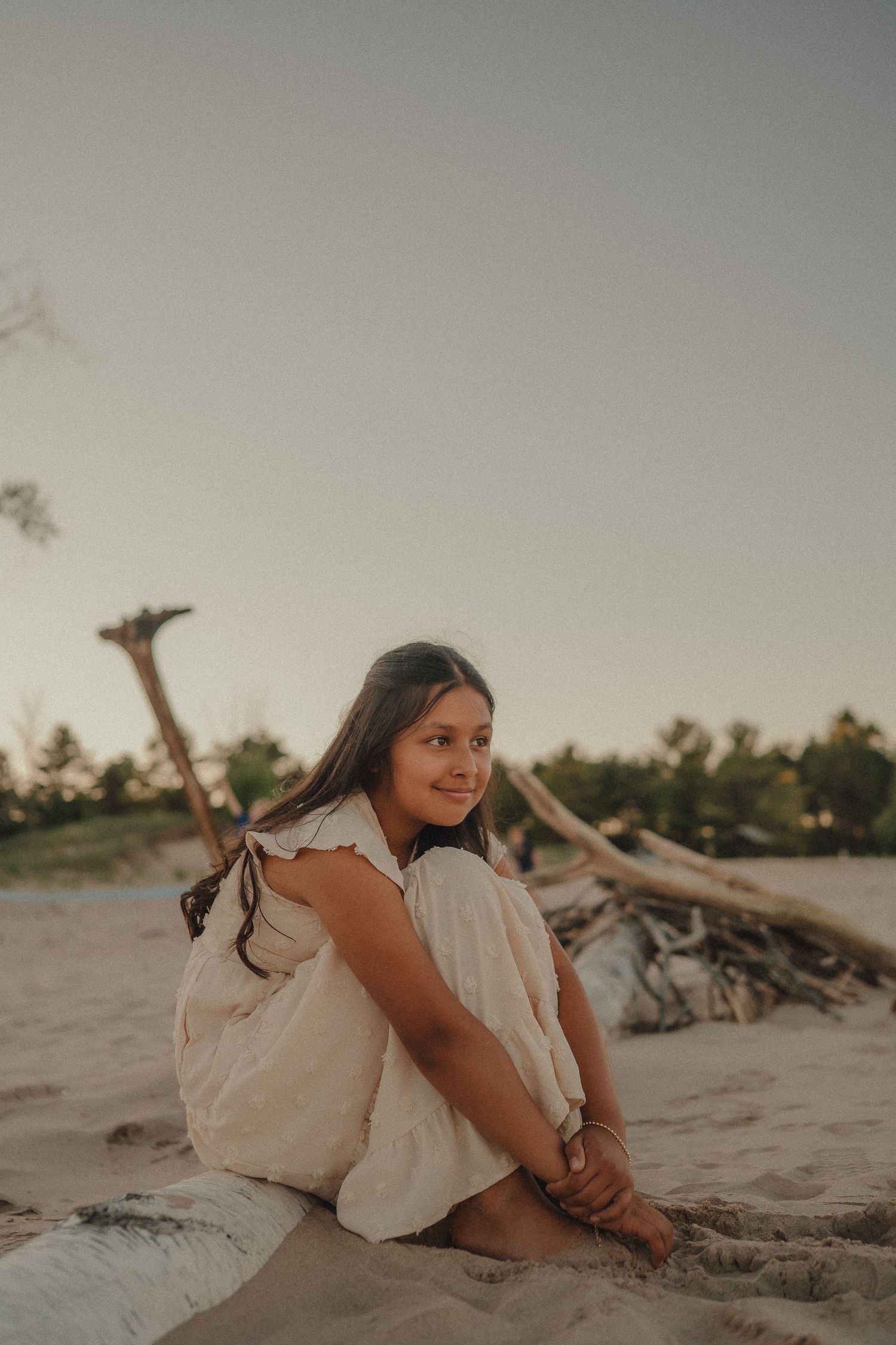 Young woman in white dress sitting on sand at beach during golden hour sunset.