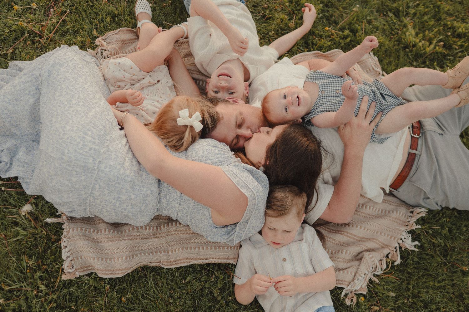 Aerial view of a mother and four young children lying on a blanket in green grass, wearing soft pastel outfits.