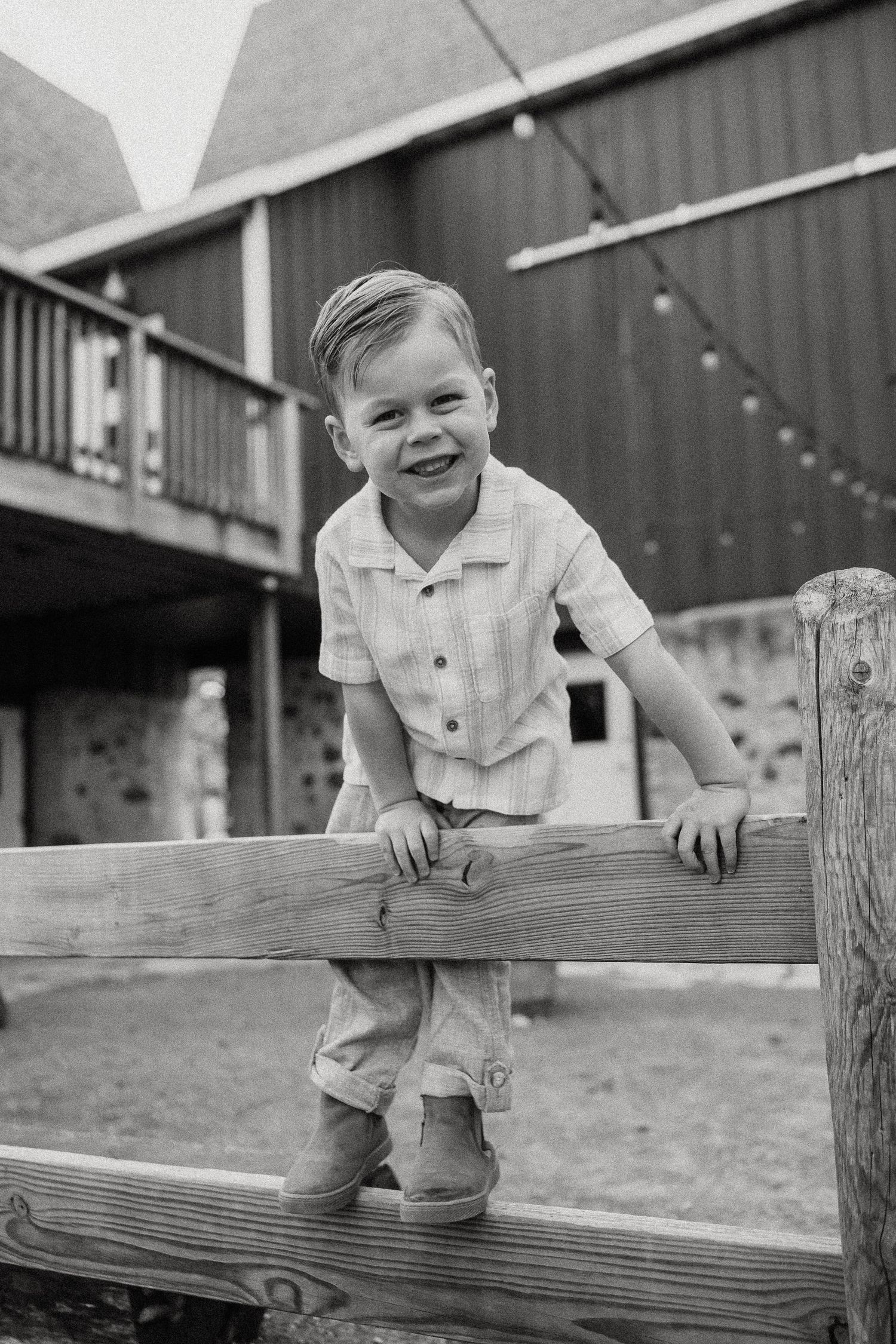 Smiling young boy in vintage clothing leaning on a wooden fence outside a red barn in a black and white photo.