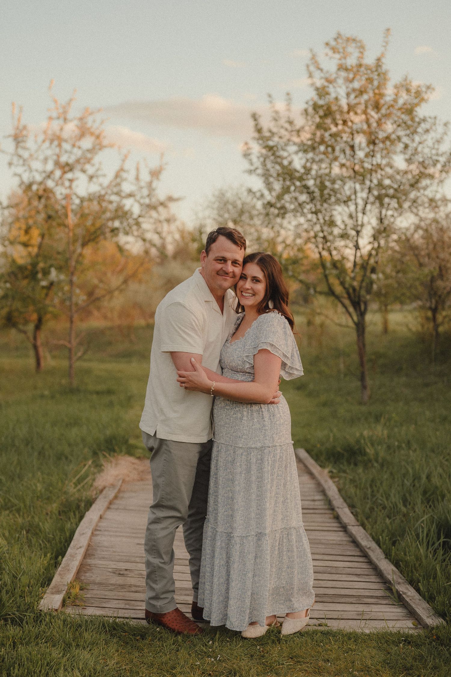 Couple embracing on a wooden bridge surrounded by lush green trees during golden hour sunset.