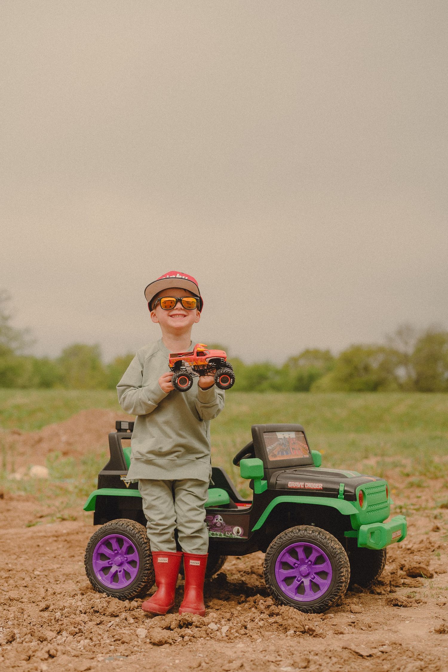 Young boy in red boots and sunglasses holding a monster truck toy beside a green ride-on jeep outdoors.