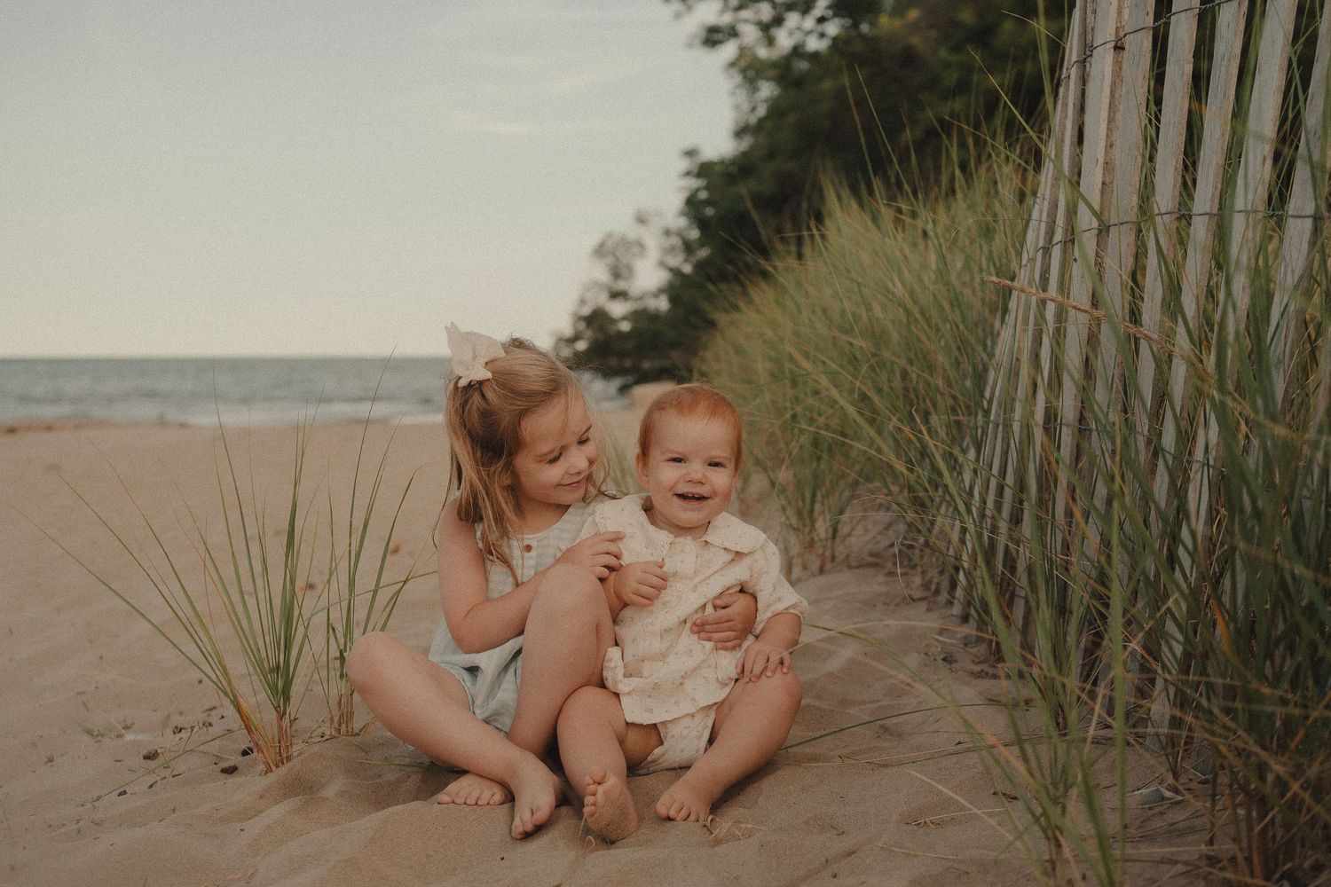 Two young sisters sit together on sandy beach dunes near grass and a fence, smiling joyfully by the ocean.