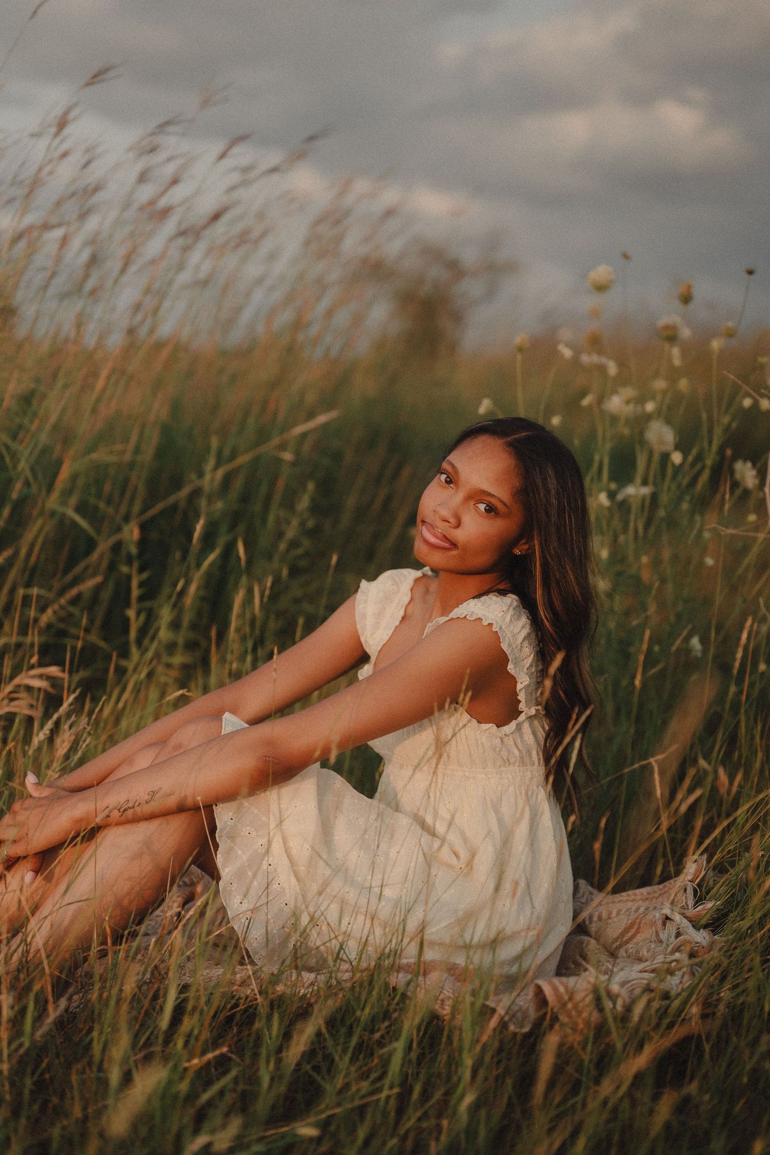 Young woman in white lace dress sits relaxed in a golden meadow of tall grass at sunset.