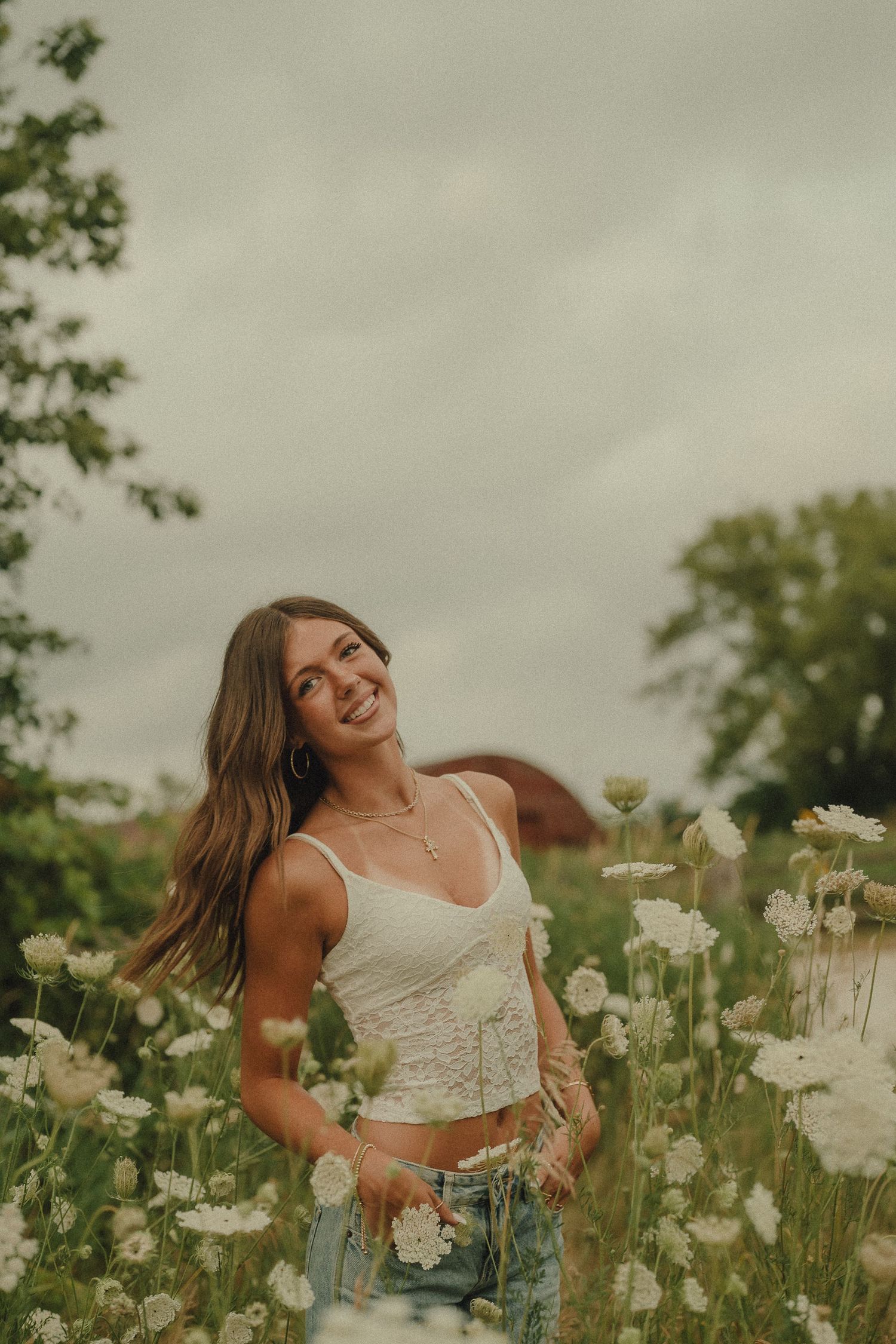 Smiling young woman in white lace crop top posing joyfully among white wildflowers in an outdoor field.