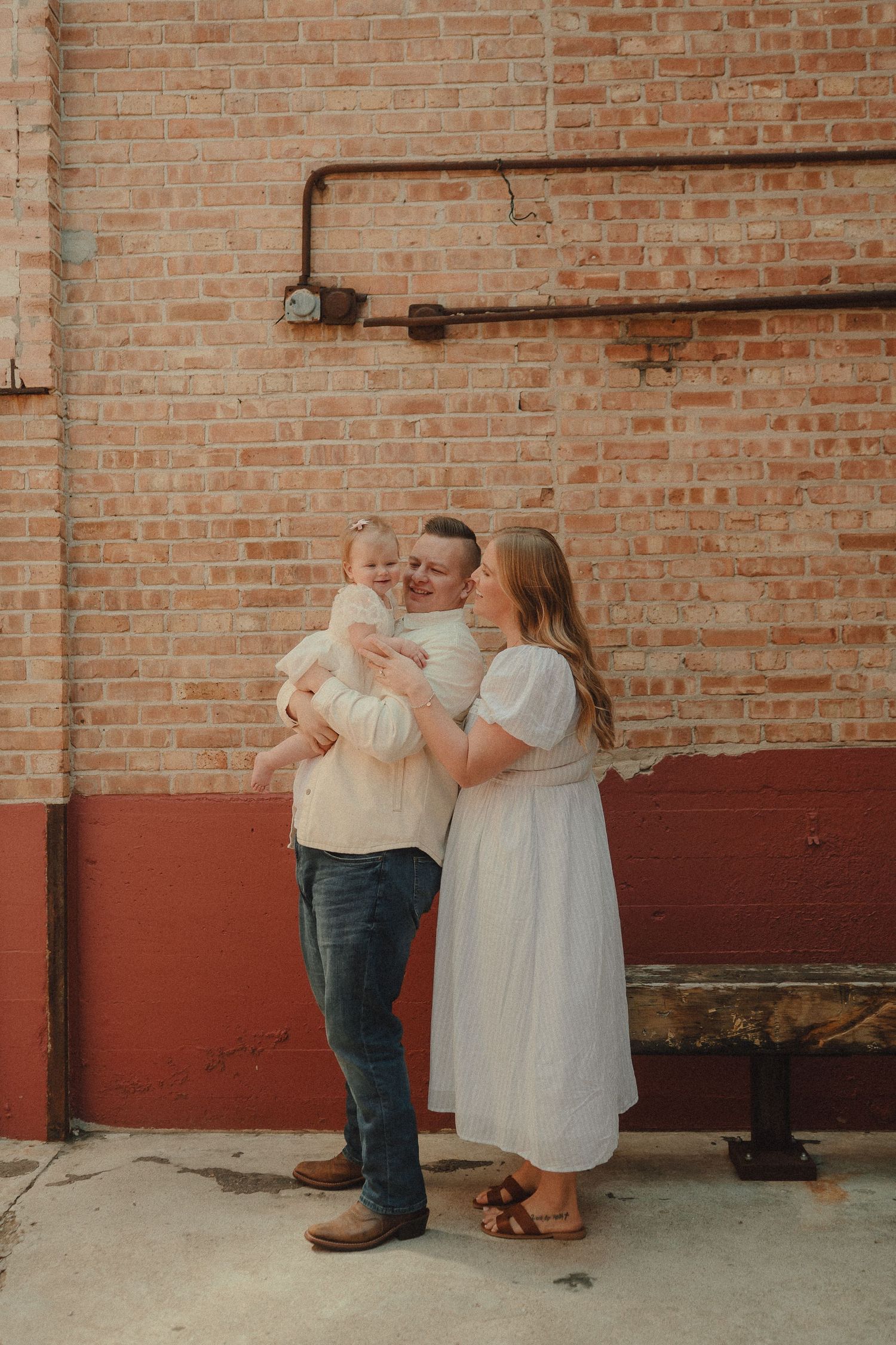 Happy family of three shares a joyful moment against a rustic brick wall during an outdoor photo session.