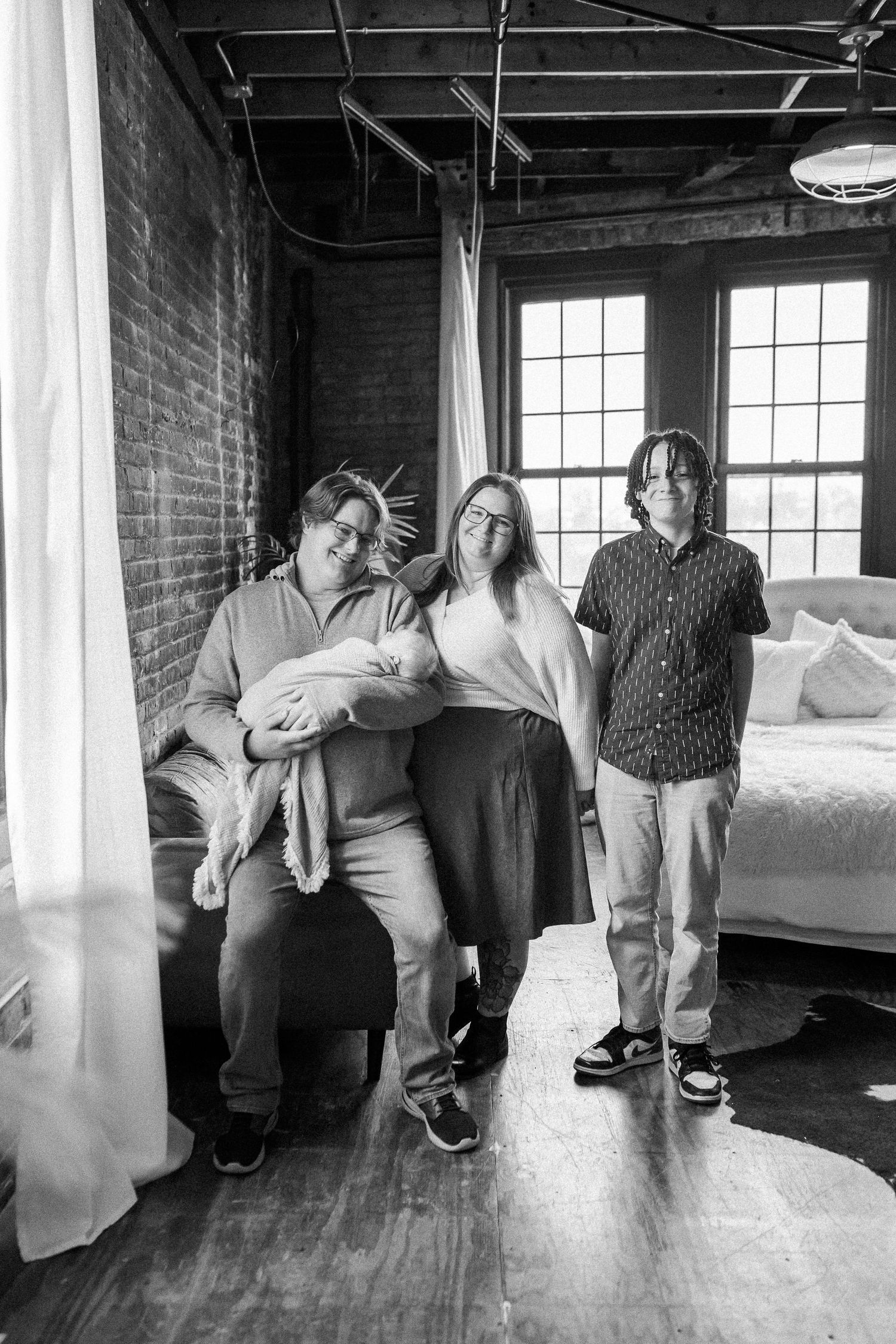 Black and white family portrait with parents and teen child holding newborn in a loft-style room.