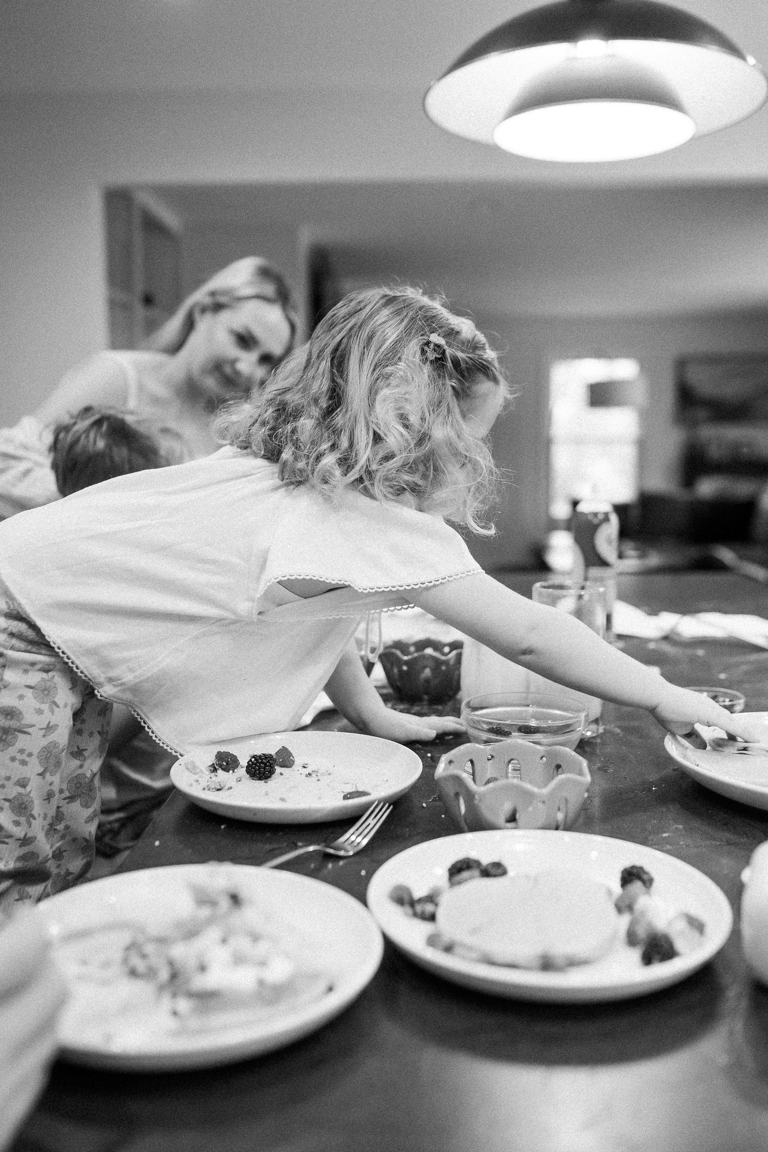 Black and white photo of a young girl reaching across a dinner table while a smiling woman watches in the background.