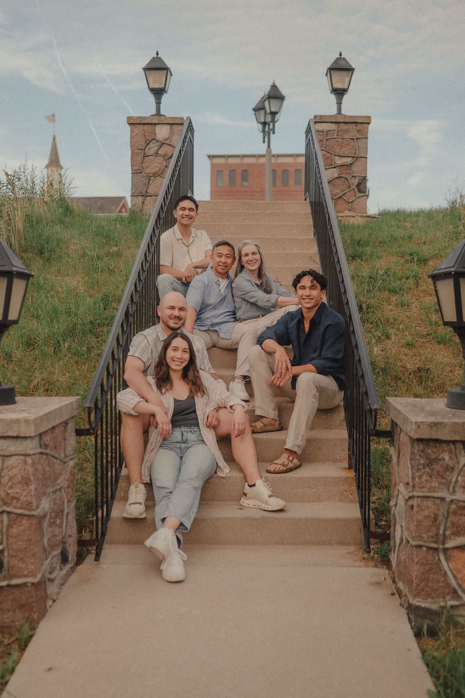 Six friends smiling and posing together on outdoor stone steps with lamp posts and brick building backdrop.