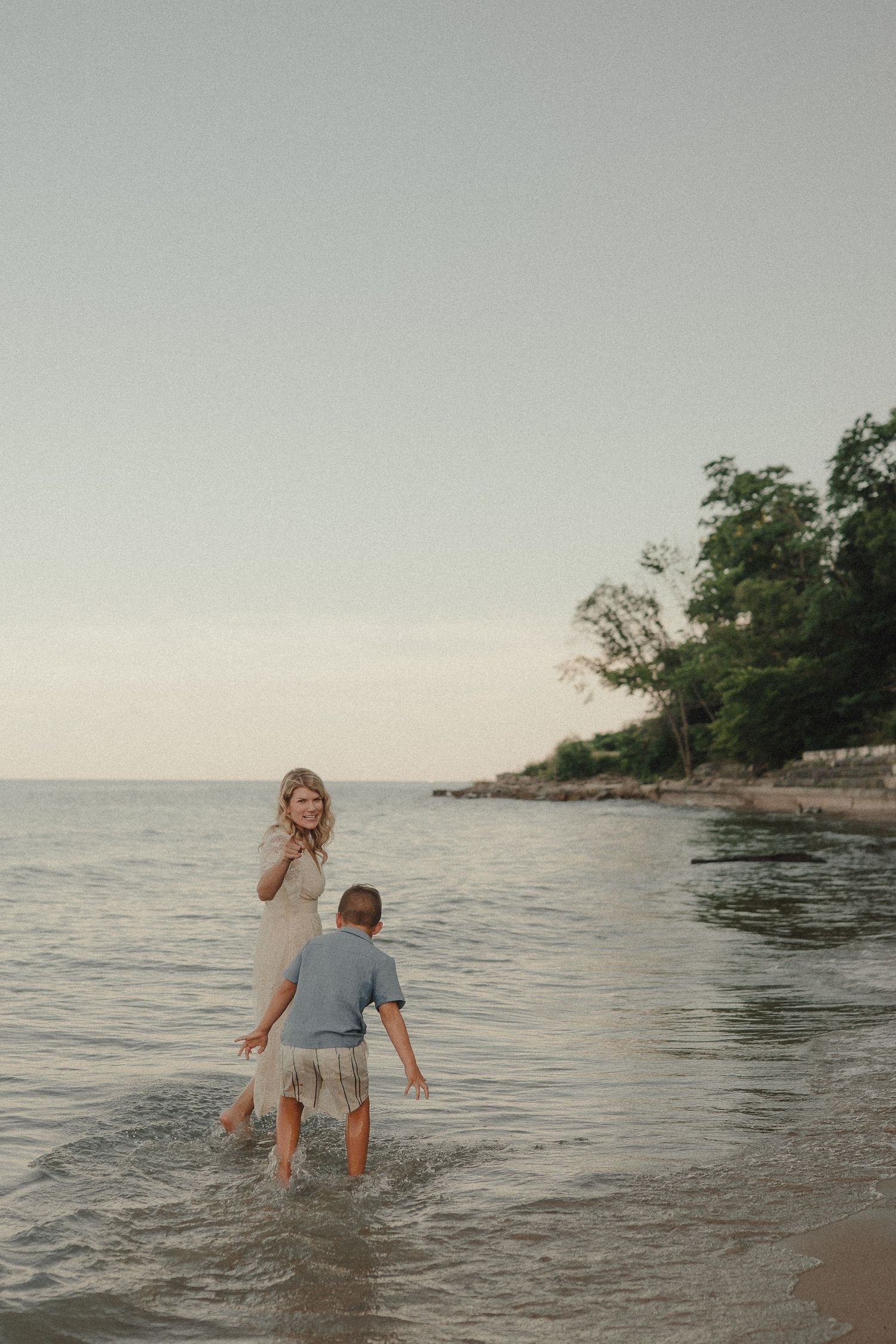 Mother in white dress playing with young son in shallow ocean waves at dusk.