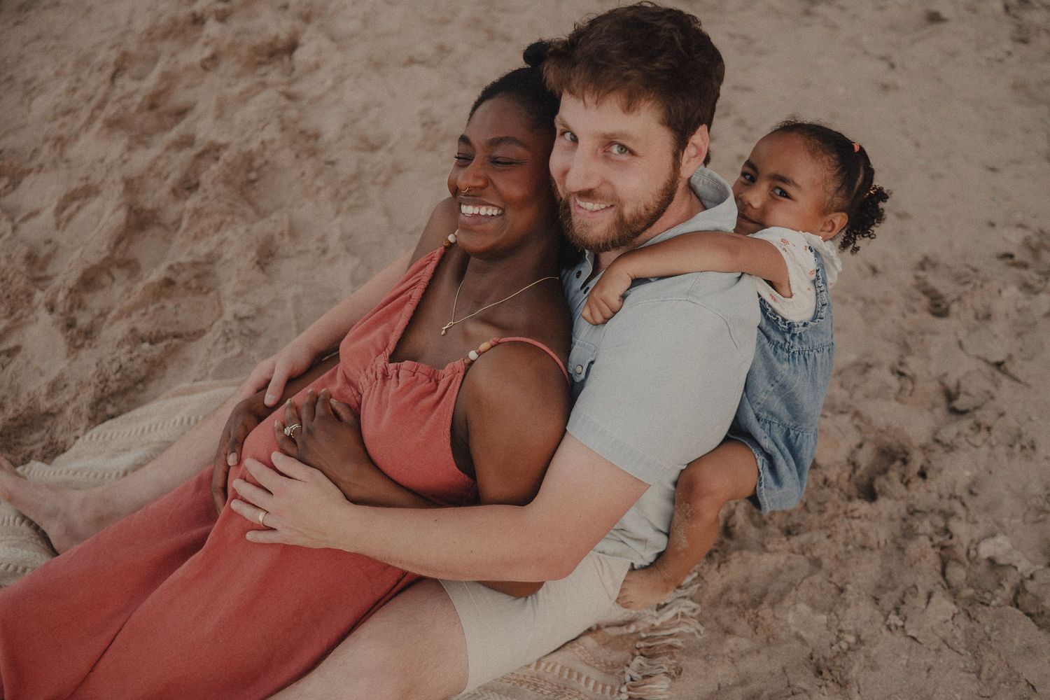 Happy interracial family relaxing together on sandy beach, parents smiling with young child outdoors.