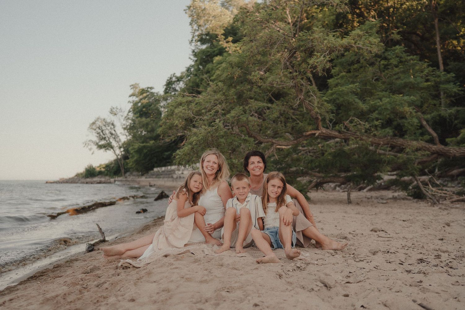 Four smiling women and girls sit together on sandy beach surrounded by lush green trees.