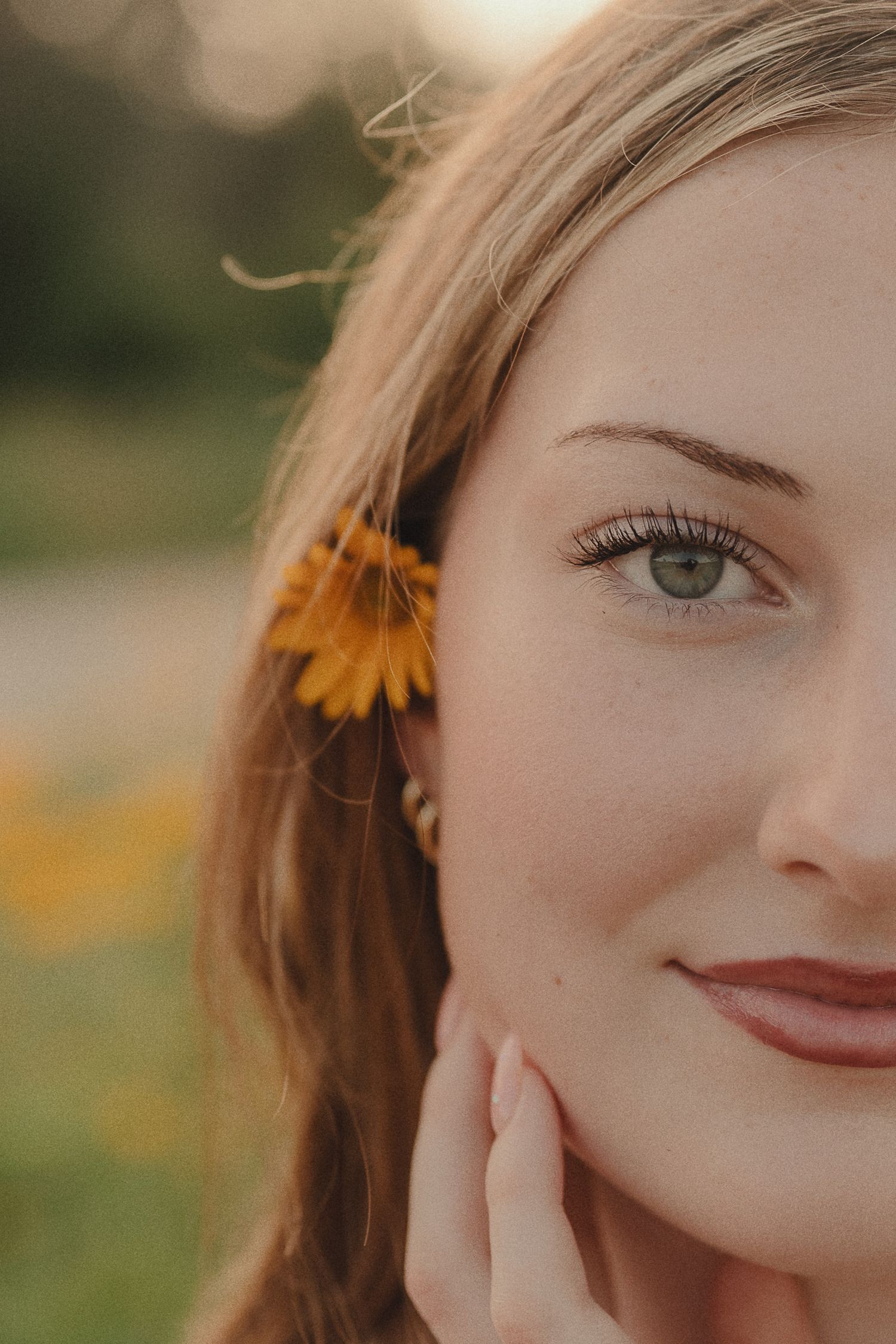 Close-up portrait of young woman with sunflower in hair and green eyes touching her face.