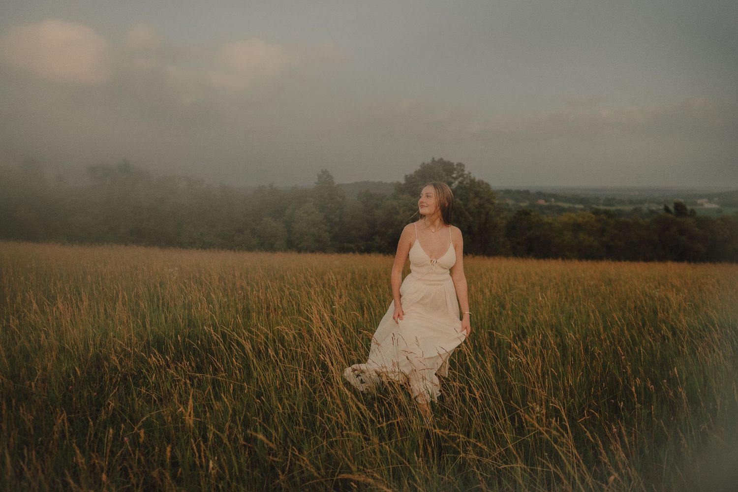 A bride in a flowing white lace gown stands in a golden field at dusk with misty hills in the background.