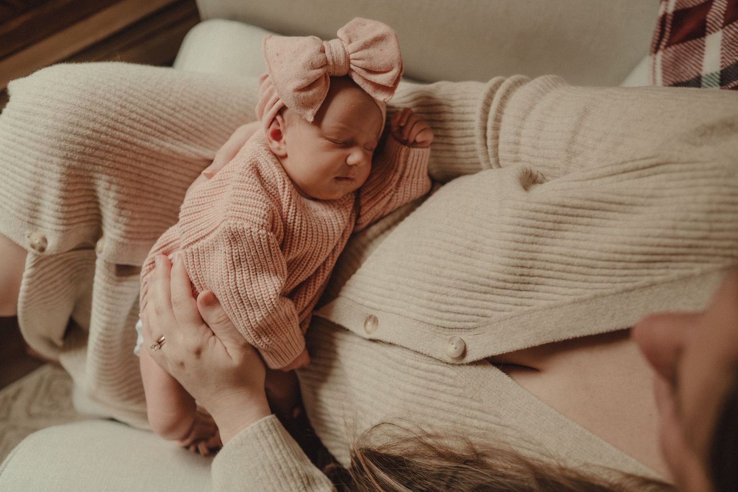 A baby in pink clothing lies on soft white bedding while reaching for a pillow.