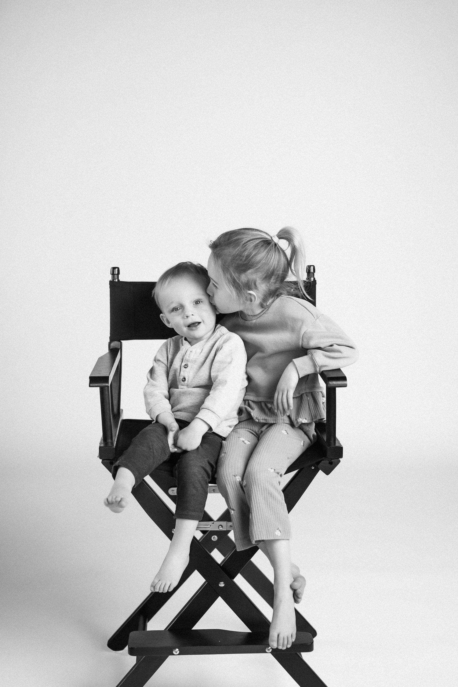 Two children share a director's chair in a playful black and white studio portrait.