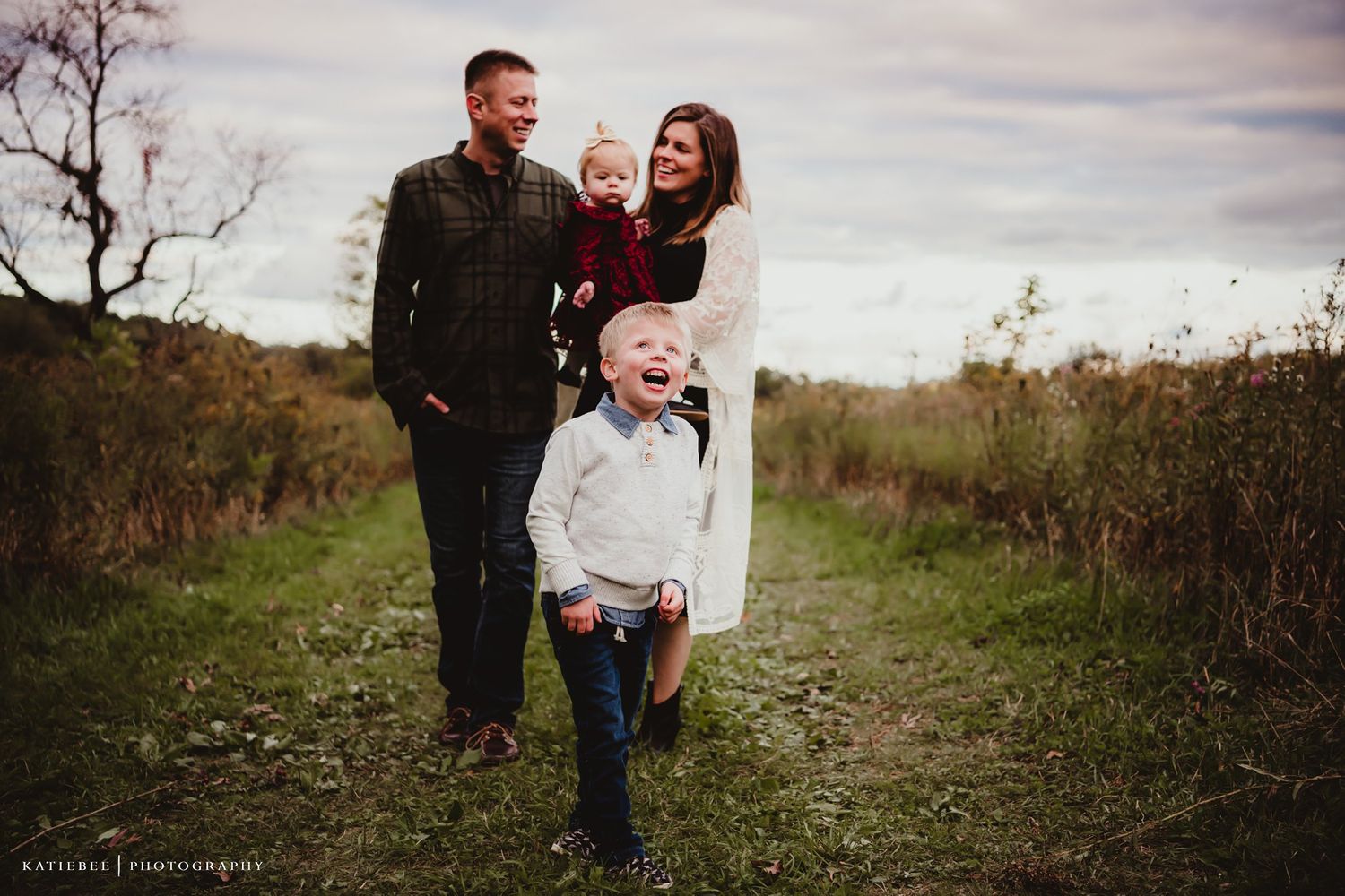 A family of four poses together on a rustic dirt path surrounded by autumn foliage.