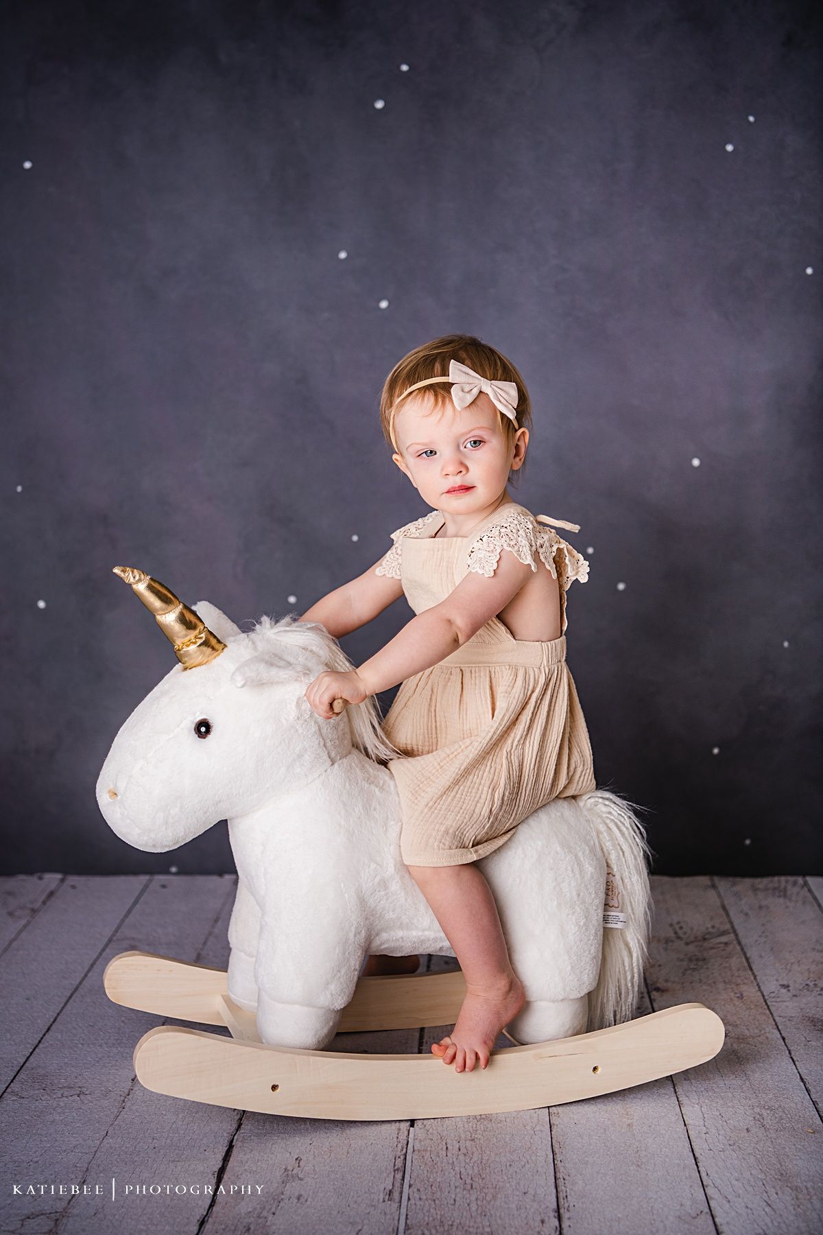 Child in beige dress sitting on white rocking unicorn toy against starry backdrop.