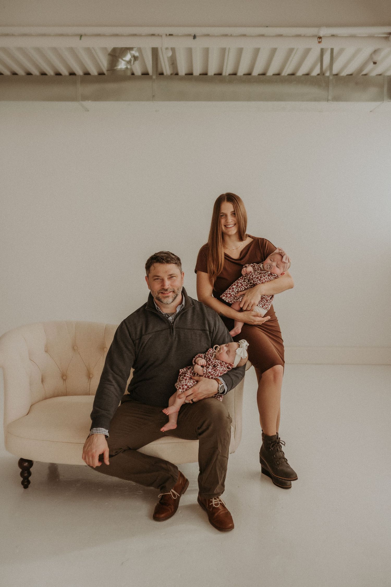 Parents with twin babies pose casually on a vintage sofa in a minimalist studio setting.