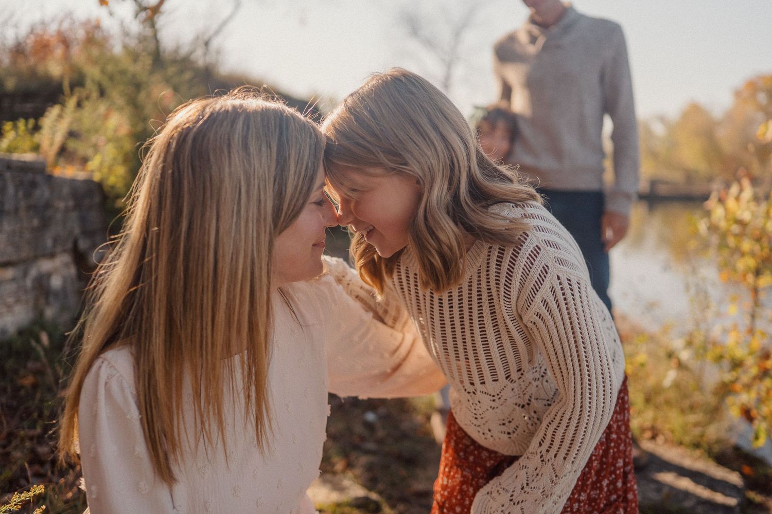 Two people share a tender moment outdoors on an autumn day with golden sunlight and fall foliage in the background.