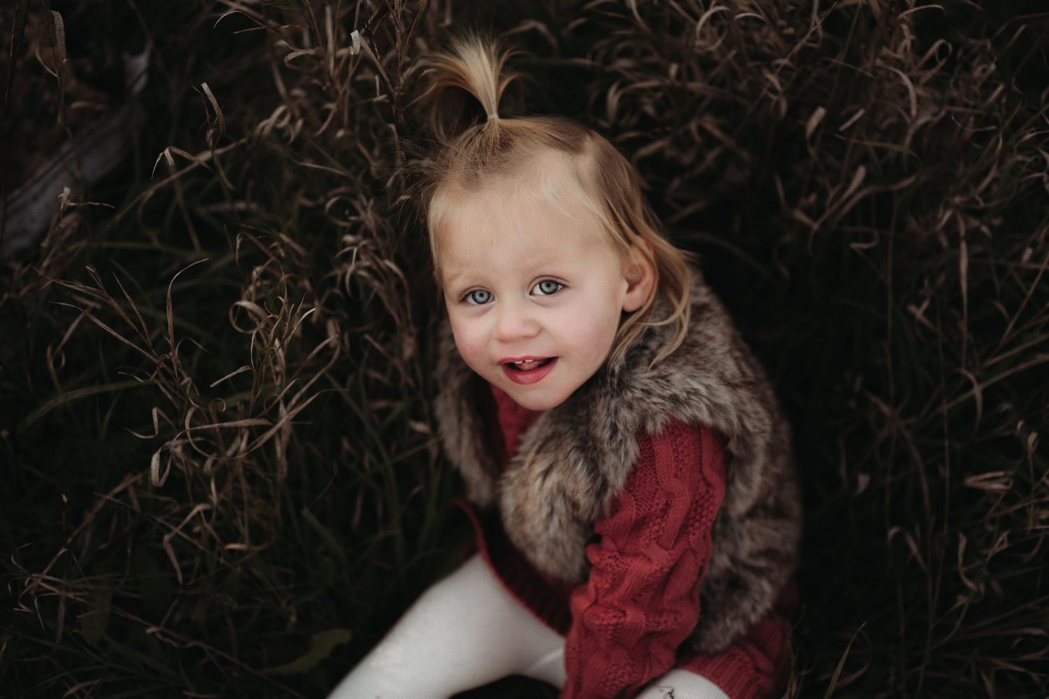 Close-up portrait of a toddler wearing a fur vest and red sweater against dark foliage.