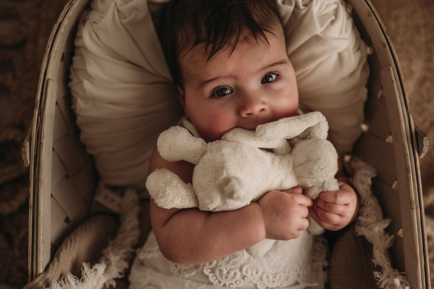 Baby cuddles soft white plush toy while resting in cream-colored bassinet.