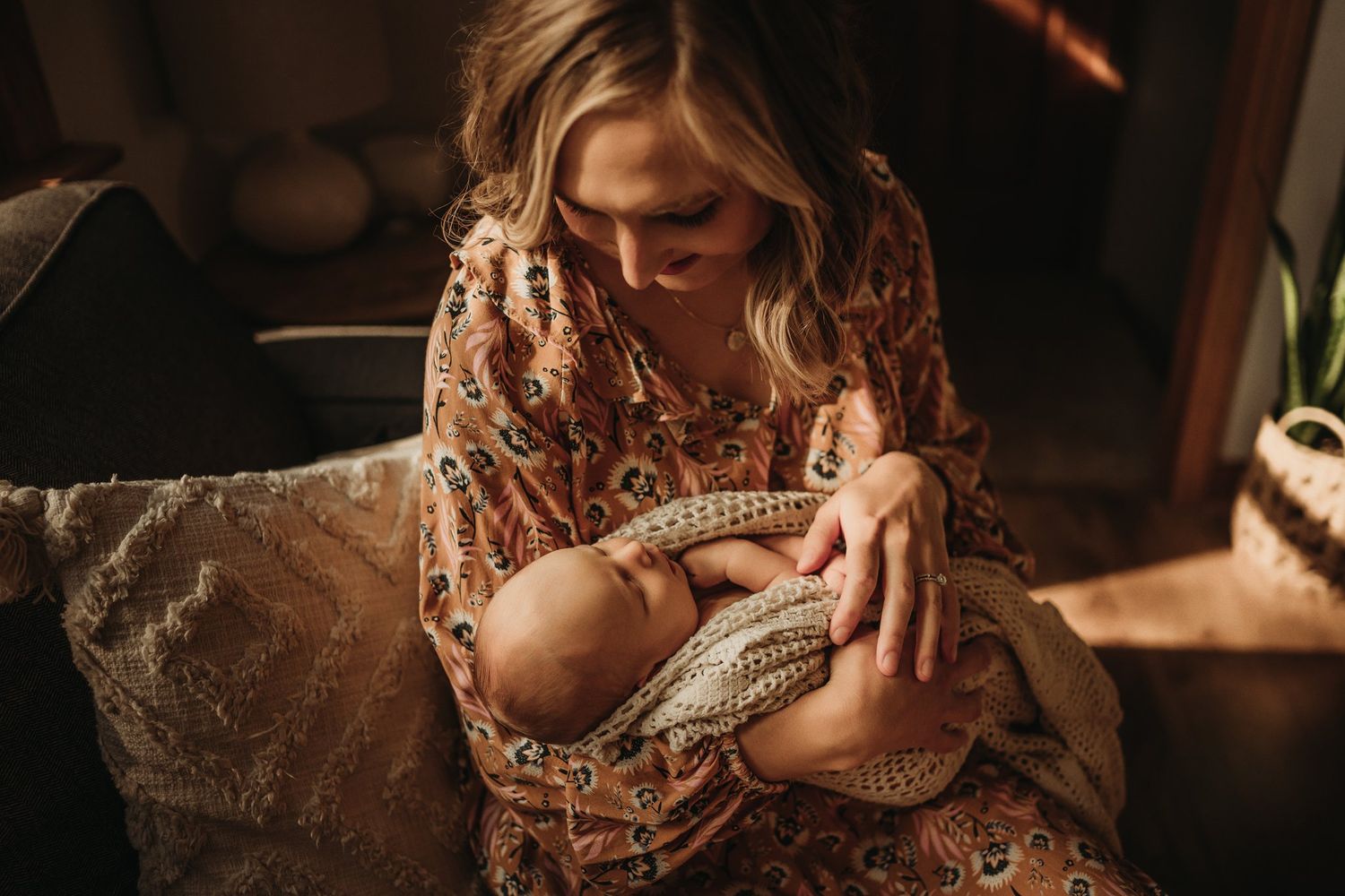 Mother wearing floral dress tenderly holds sleeping newborn wrapped in knit blanket.