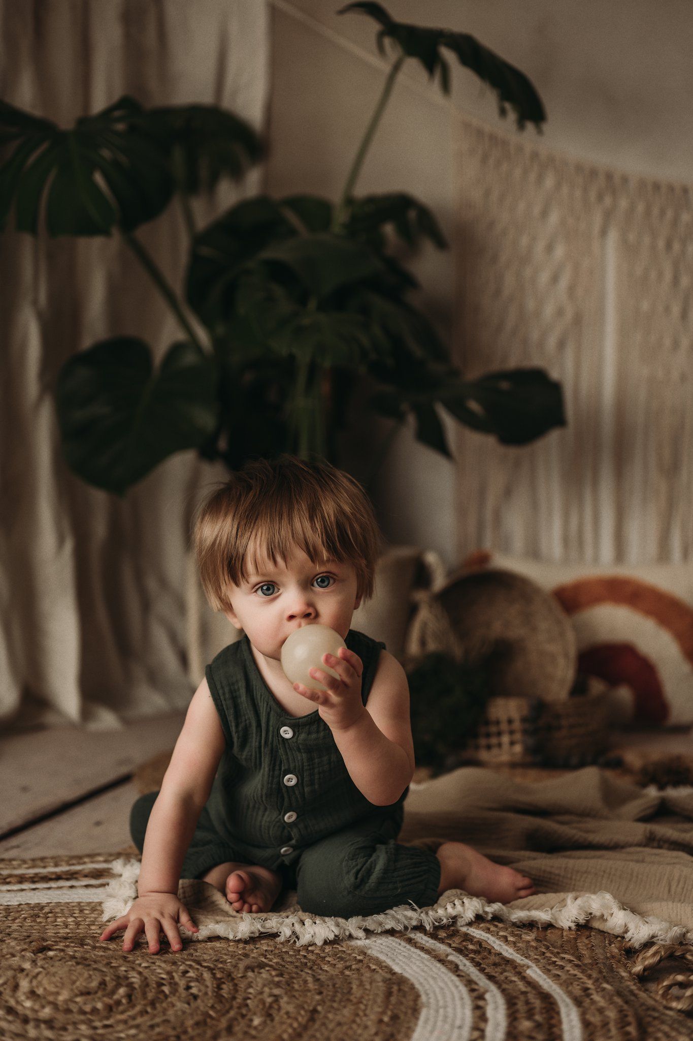 Small child in dark green overalls sits on rug with monstera plant and macrame backdrop.