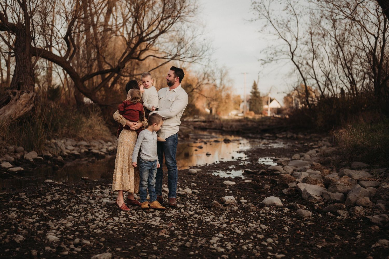 Family portrait session on rocky riverbed with bare trees and golden sunset light in background.