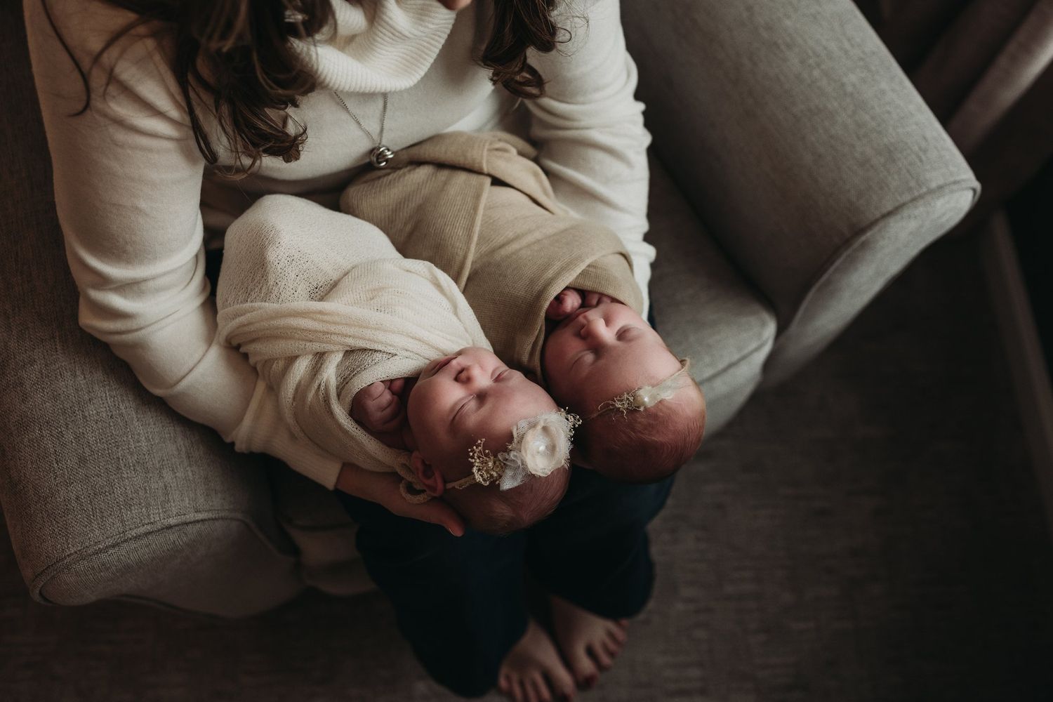 Parent cradling two infants on a gray couch in intimate lifestyle portrait.