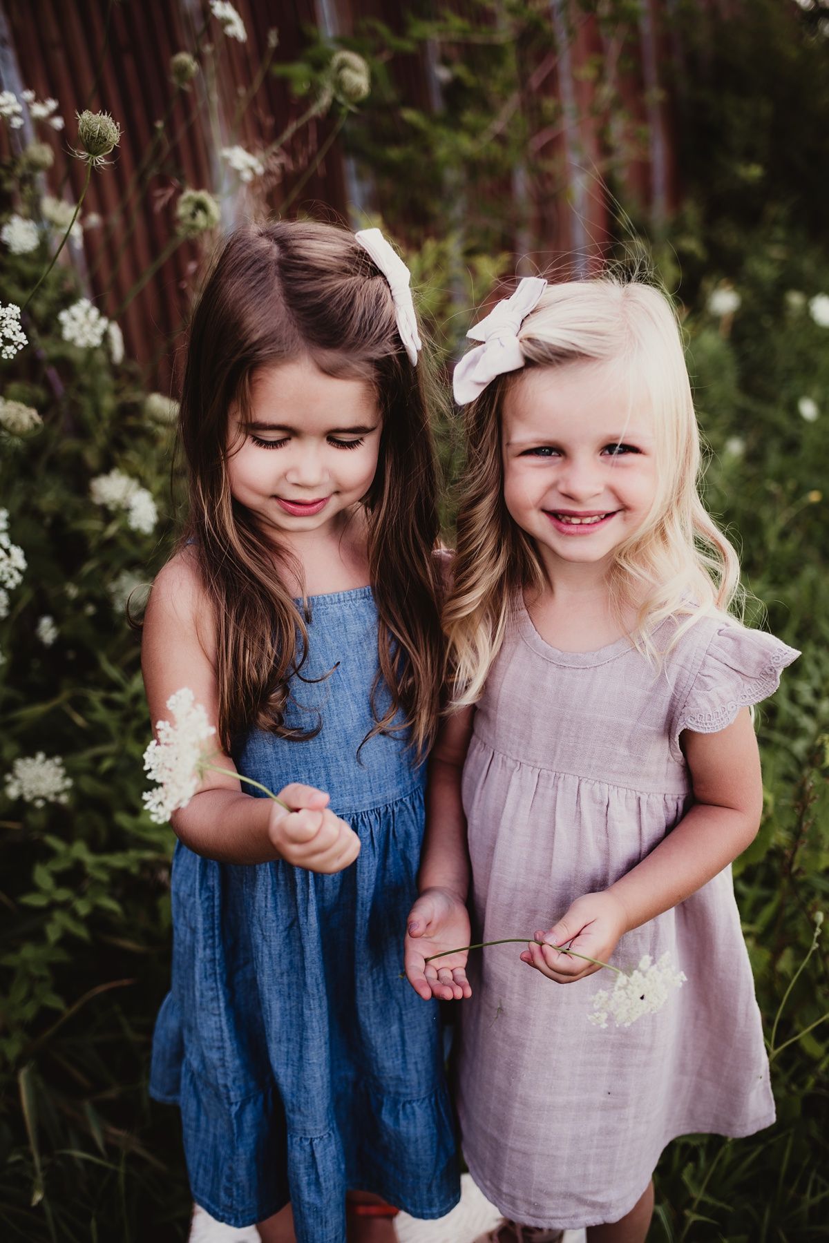 Two young girls in summer dresses stand together in a garden with wildflowers.