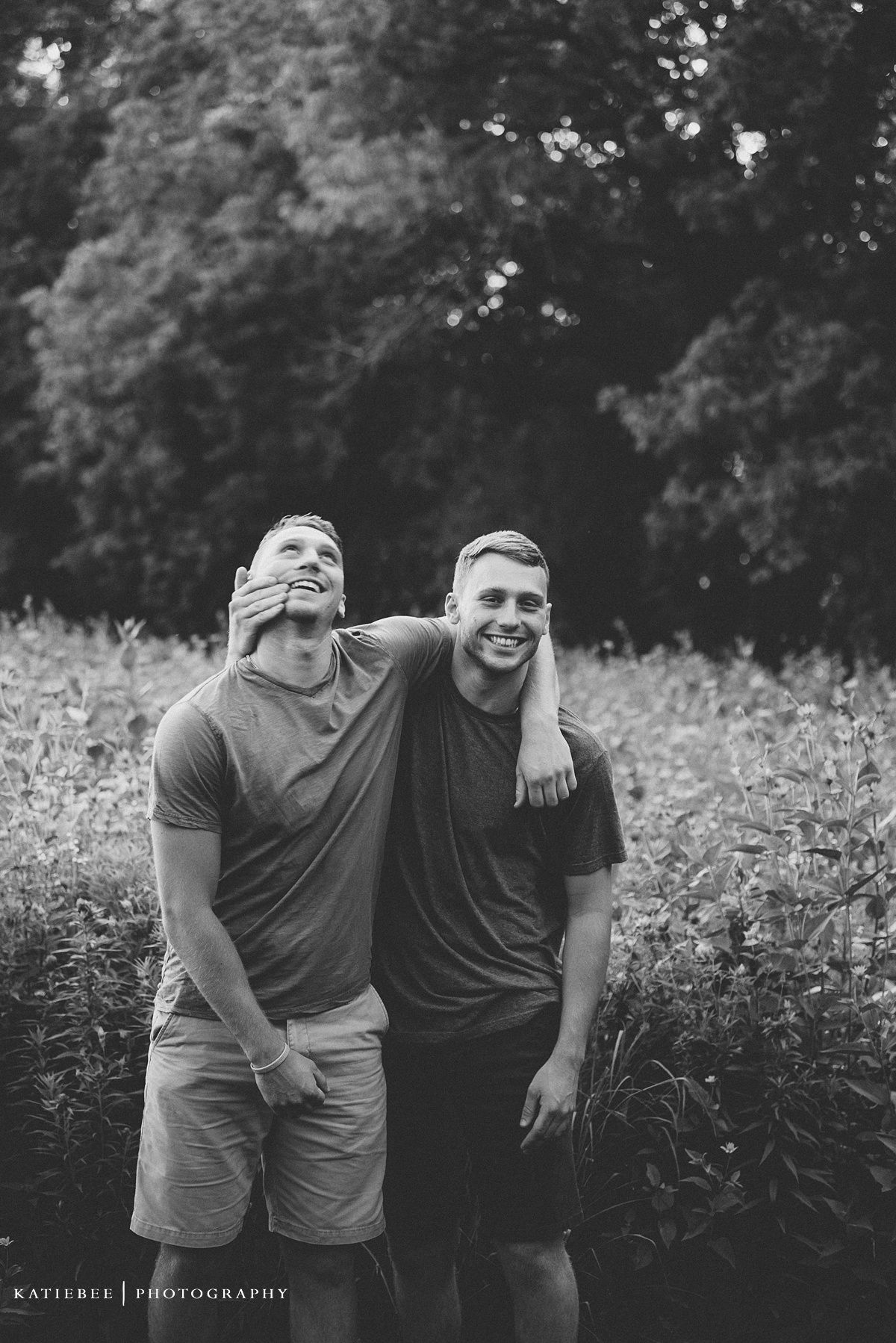 Black and white portrait of two people laughing and embracing outdoors in a wildflower field.