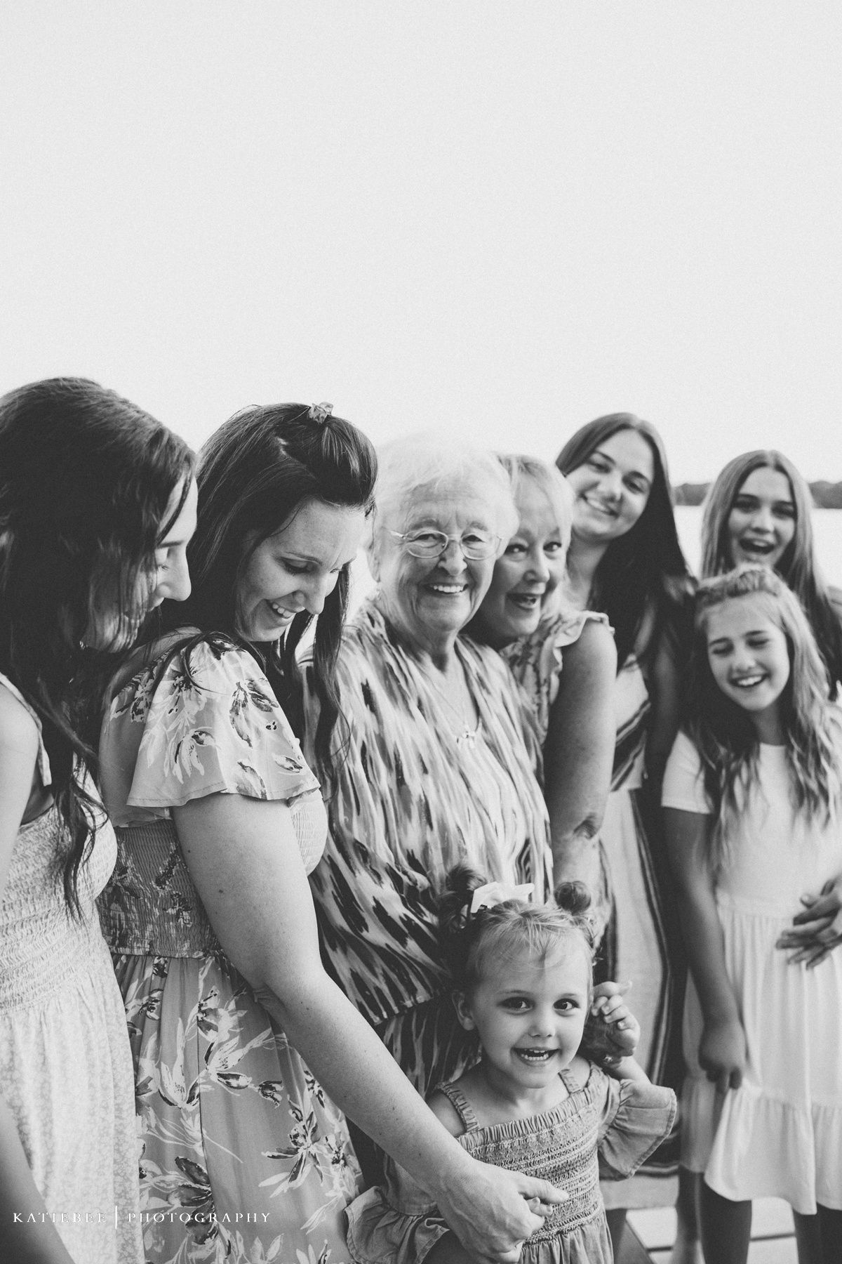 Multi-generational family in light summer dresses gather for a black and white portrait by the water.