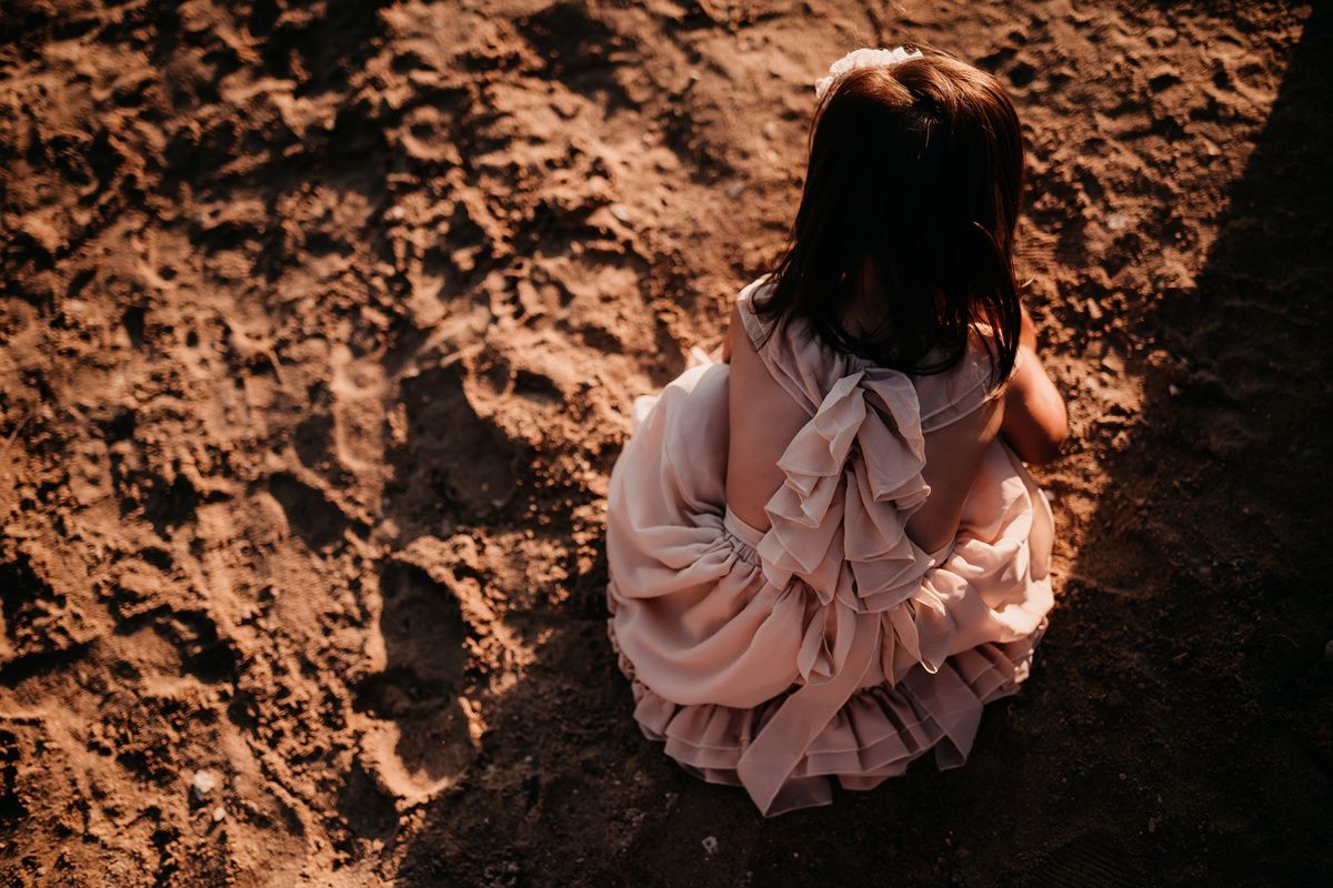 Back view of someone in a pink ruffled dress sitting on reddish-brown earth.